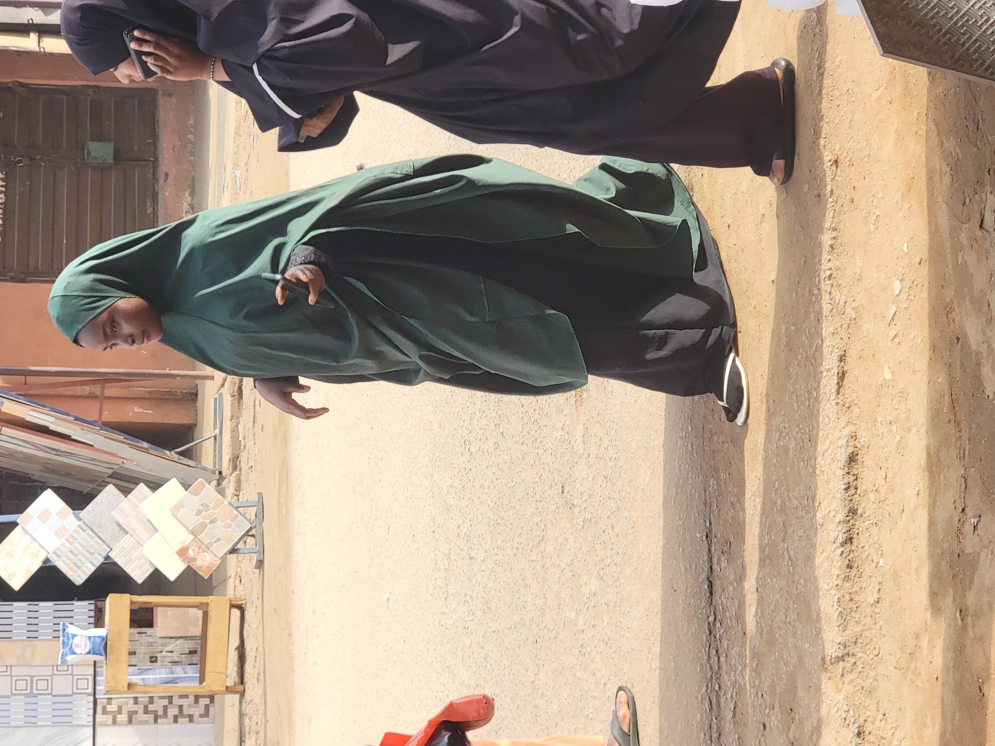 Two women are standing on a paved street in Maiburiji, Nigeria, next to a display of ceramic tiles. The woman closest to the camera is wearing a dark green hijab and a black garment underneath. Her right hand is raised. The other woman is wearing a black abaya and is holding a mobile phone to her ear. A building with a brown door is in the background. The ground is a combination of concrete and dirt. A red object is visible at the bottom of the image, near the street.