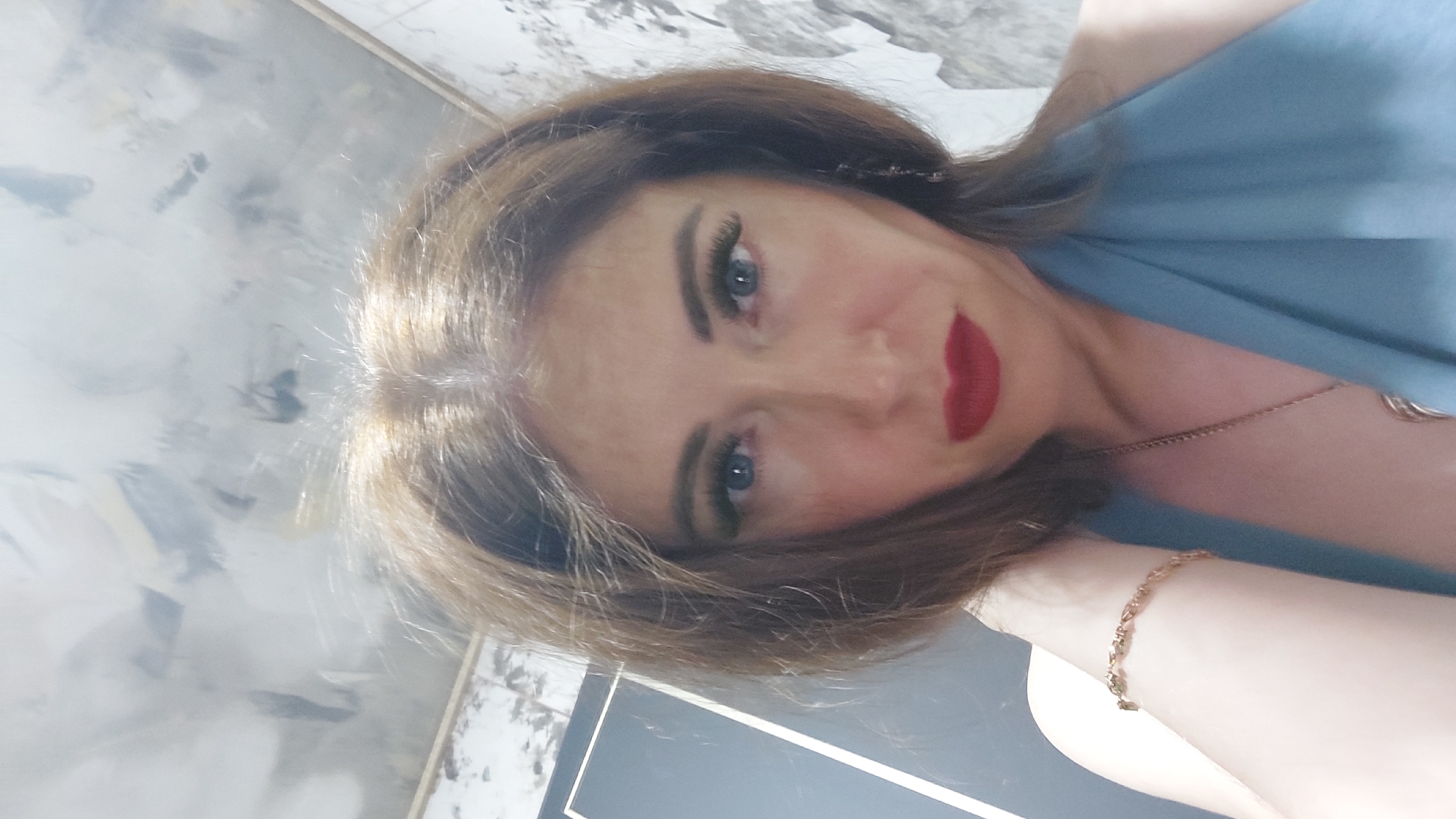 The image is a close-up, selfie-style shot of a woman with brown hair, blue eyes, and red lipstick. She is wearing a blue garment and a gold bracelet. The background appears to be an indoor setting with patterned wallpaper and a gray door or frame. There is no visual information in the image to suggest it is located in the city of Komsomolsk-on-Amur, Russia.