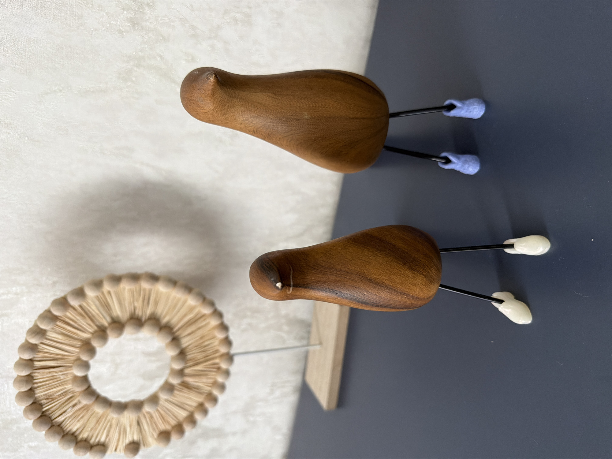 The image shows two wooden bird figurines with thin black legs and light-colored feet, standing on a dark blue surface. The figurine on the left has white feet, while the figurine on the right has light blue feet. Behind the figurines, there is a circular, fan-like wooden decoration with beads around its perimeter, supported by a thin white rod and a wooden base. The background is a textured white wall. The scene is indoors, with soft lighting. There is no indication of the city of Novosaratovka, Russia, or any elements suggesting a specific geographic location.