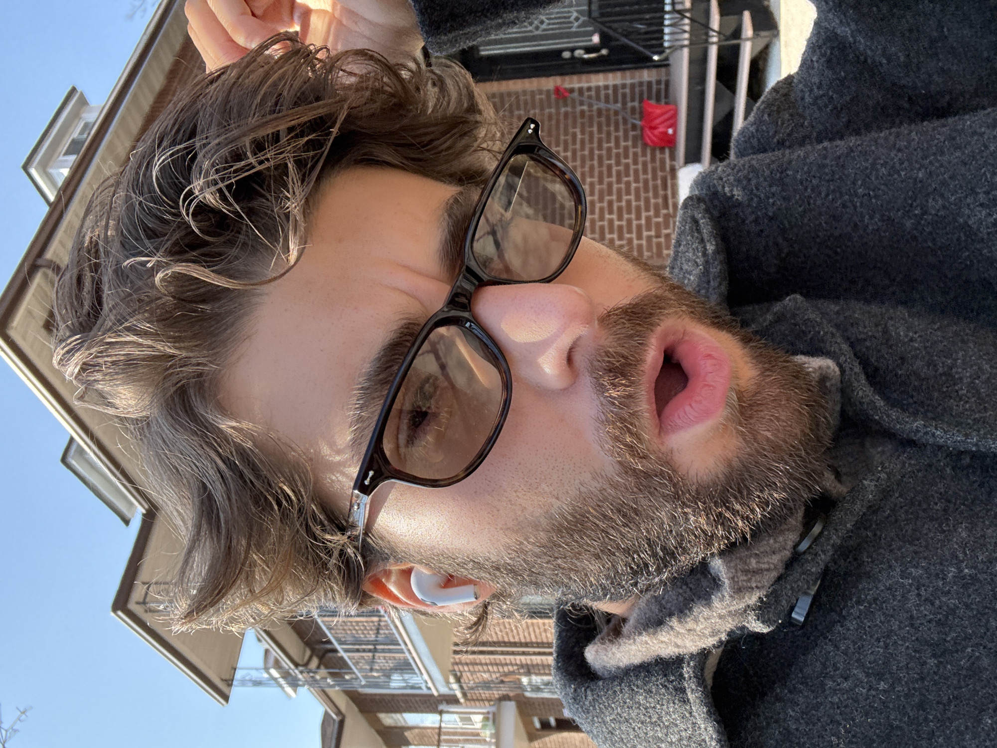 A close-up, slightly angled, outdoor photograph features a Caucasian man with wavy brown hair and a beard, wearing dark-rimmed glasses and a gray wool coat. He has an open mouth and appears to be looking upwards. A white wireless earbud is visible in his left ear.
In the background, indistinct building facades and a blue sky are visible, suggesting an urban environment. The architecture suggests the location might be in Montreal, Canada, although this cannot be definitively confirmed. A red bag hanging on a brick wall is also visible.