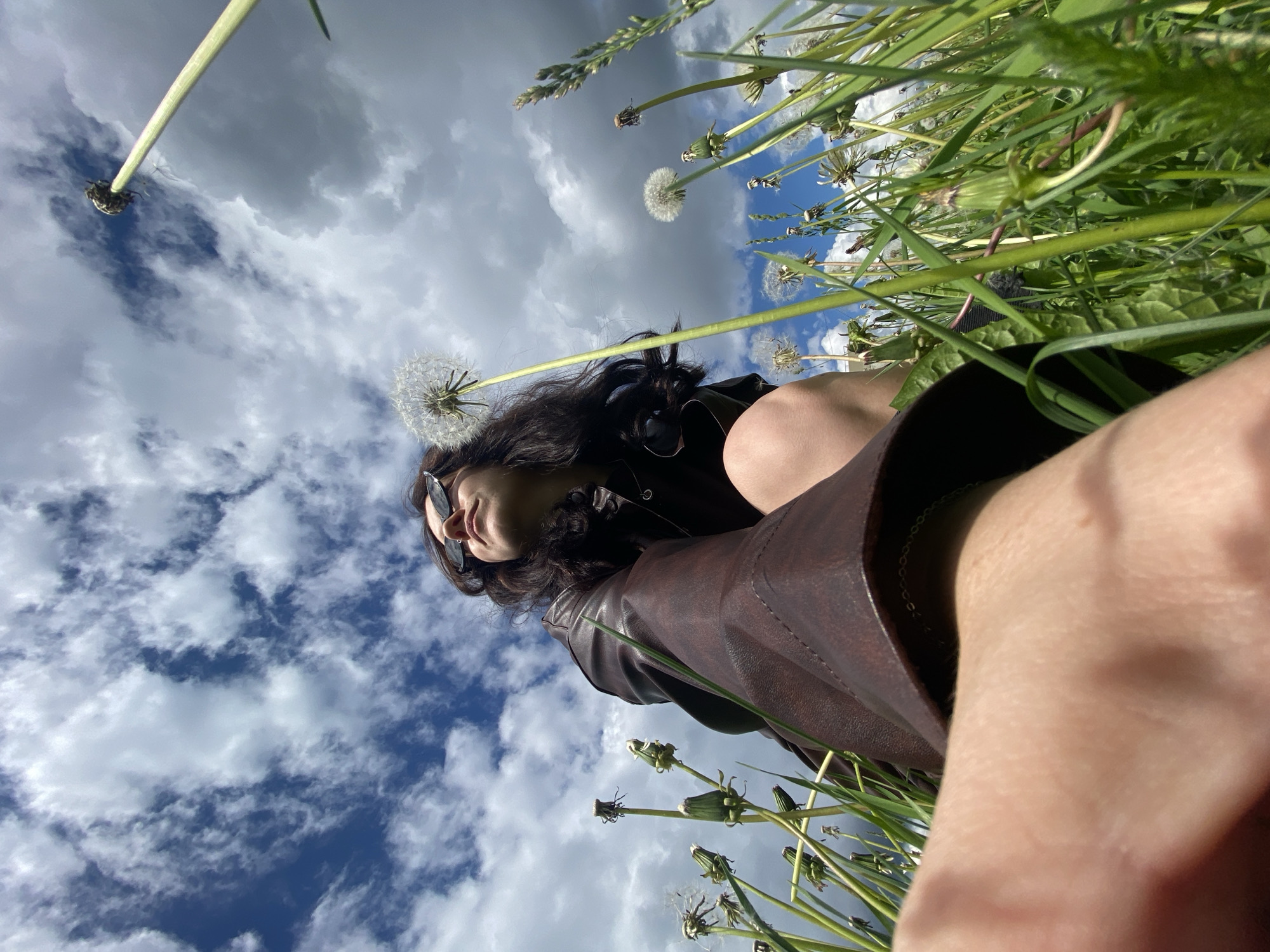 A low-angle, wide shot depicts a woman lying in a grassy field with dandelions and the sky above. She is wearing sunglasses and a brown leather jacket, and her dark hair is loose. Some dandelions are in bloom, while others have gone to seed, their white, fluffy heads contrasting against the blue sky with scattered clouds. The composition is unusual, giving the impression of the woman lying on the earth, looking up at the sky. The location appears to be in a natural setting, with the specific city mentioned of Марковский, Russia, being of note.
