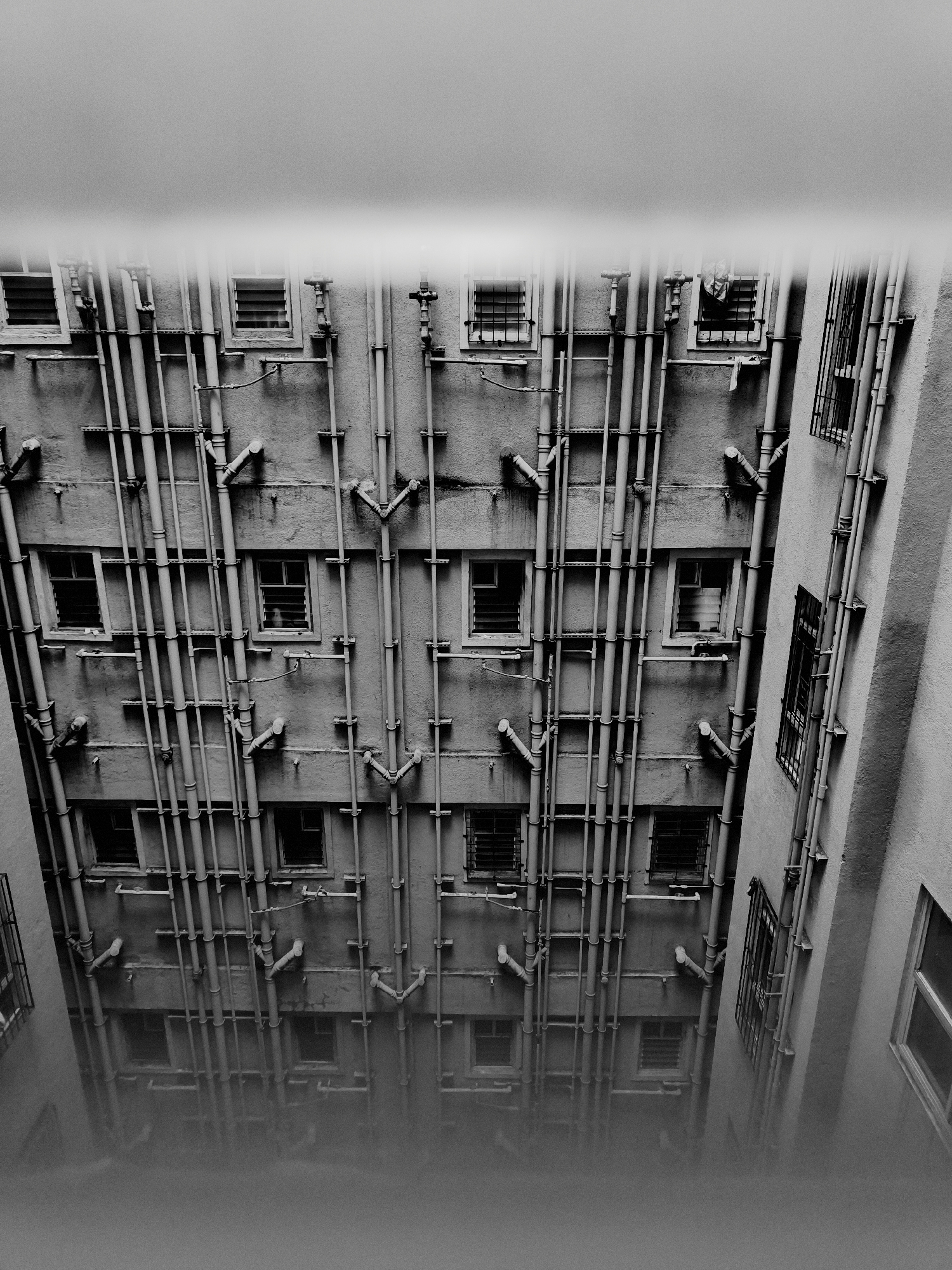 The image captures an upward-facing view of the exterior of buildings, characterized by a dense network of pipes and several windows. The architecture appears to be in an urban setting, possibly an alleyway or courtyard, with a high density of vertical and horizontal pipework running along the walls. The windows are small and some are fitted with louvers or grates. The lighting suggests it's daytime, but the overall atmosphere is somewhat stark and industrial due to the monochrome palette and the prominent pipes. The bottom portion of the image is obscured by a grey, out-of-focus haze, which could be fog, smoke, or a stylistic photographic effect. There are no people visible in the image. The location is identified as Bavdhan, India.
