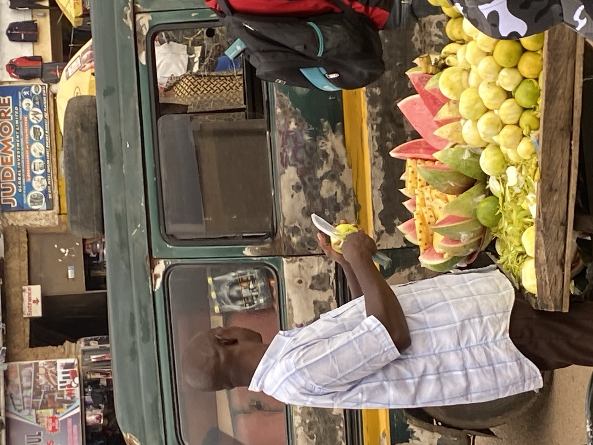 In Bukuru, Nigeria, a bald Black man in a blue and white checkered shirt is standing next to a produce stand laden with fruits. He is holding a knife and a yellow fruit, appearing to be preparing it for sale. Behind him, a green, weathered vehicle is parked. The fruit stand displays a variety of fruits including watermelon slices, pineapples, limes, and what appear to be lemons or limes. The background shows a marketplace with shops, signage, and other people.