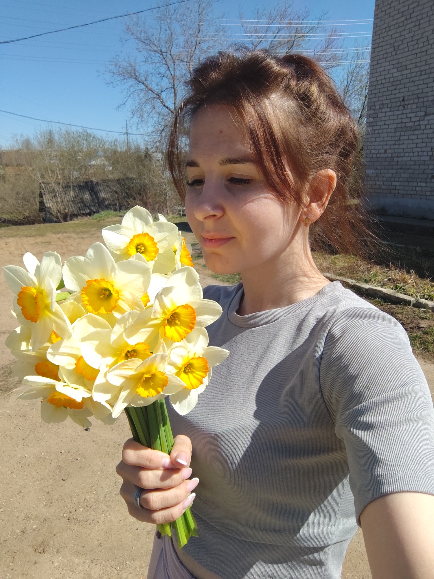 A woman holds a bouquet of white and yellow daffodils in an outdoor setting in Фоки, Russia. She is wearing a gray shirt, and her hair is styled in an updo with bangs. The background shows a dirt area, trees, a building made of light gray bricks, and a clear blue sky.