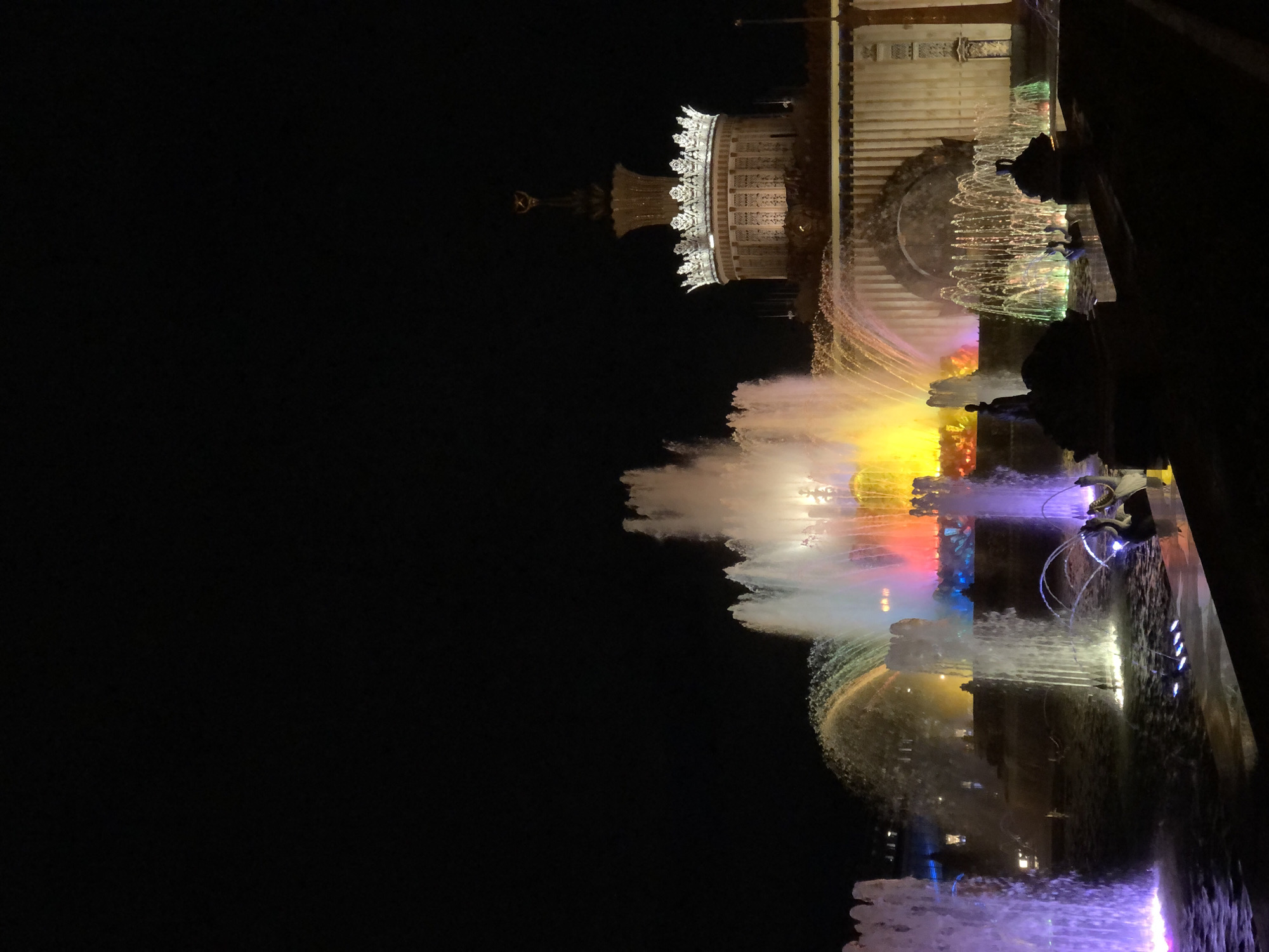 The image depicts the Friendship of Nations Fountain at Exhibition of Achievements of National Economy (VDNKh) in Moscow, Russia, at night. The fountain is illuminated with various colors, including white, yellow, red, blue, and purple. The fountain's water jets reach high into the air, creating a dynamic display. In the background, a building with white architectural details is visible, also illuminated. The night sky is dark and provides a stark contrast to the bright fountain and building.