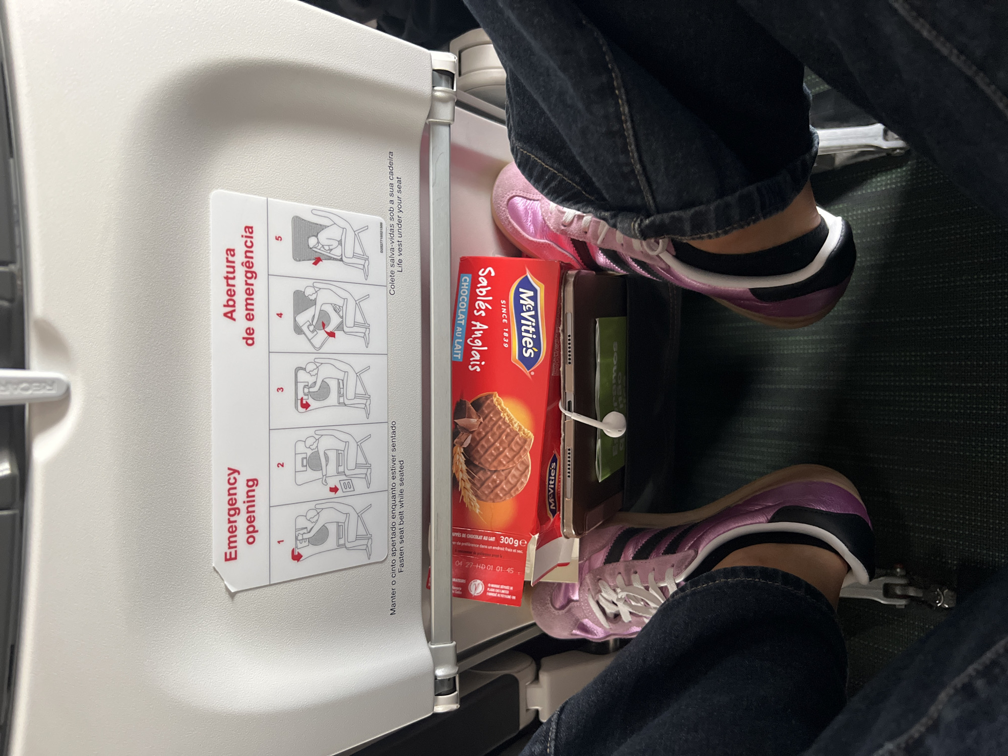 The image shows the leg and shoe of a person on an airplane. The person is wearing dark blue jeans and pink and black athletic shoes. In front of the person's legs is a tray table, on which a box of McVitie's chocolate biscuits and a tablet are placed. An emergency instruction card is visible on the back of the seat in front of the person. The location is the cabin of an airplane, in Paray-Vieille-Poste, France.