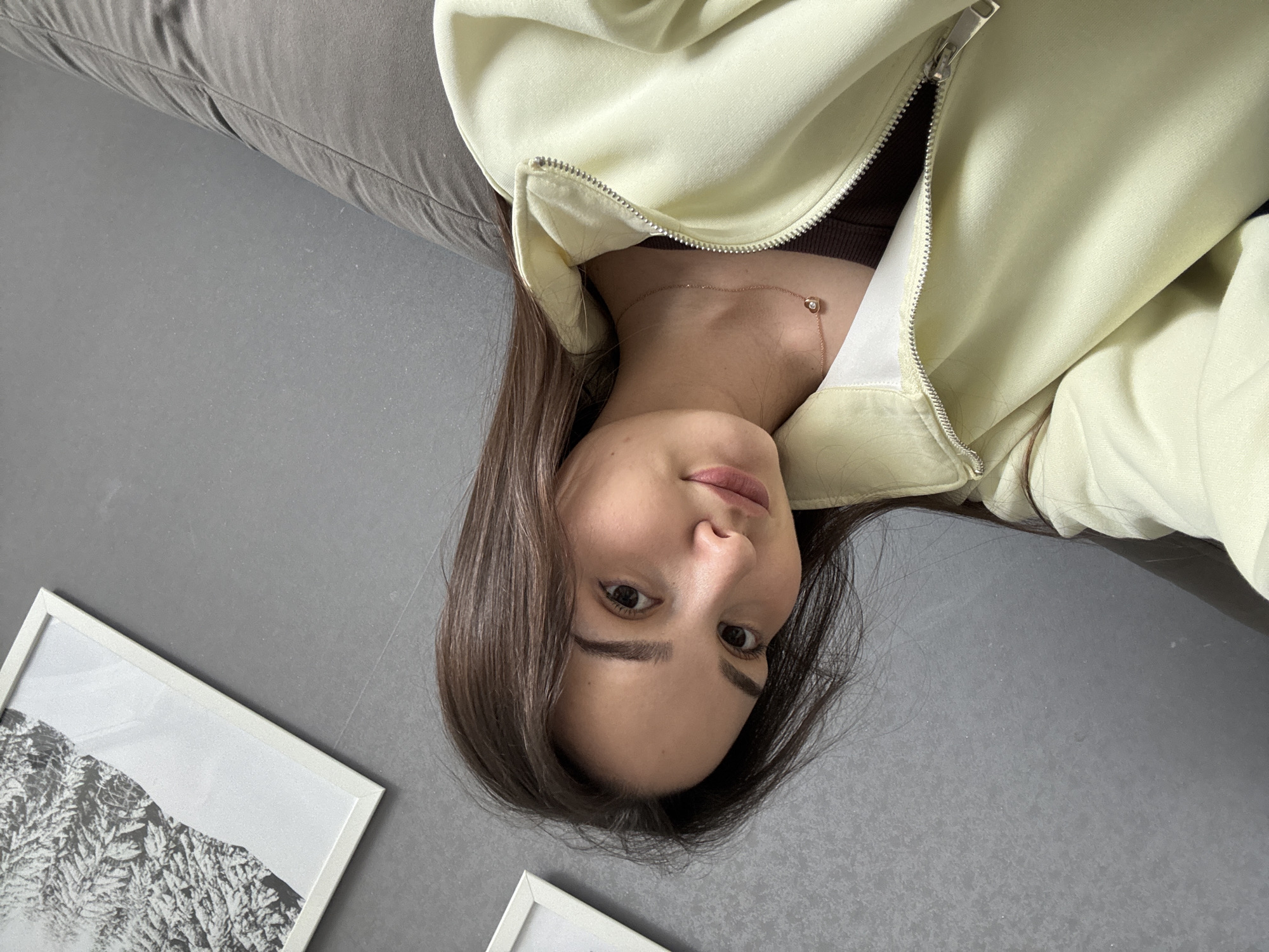 A woman is lying down, photographed from above. She has brown hair and is wearing a brown top with a light green zippered jacket. She also wears a gold necklace with a small pendant. The woman is lying on a gray surface near a gray sofa. In the lower left corner, there are two framed pictures. The location appears to be indoors. There is no information available to determine if this image was taken in Chaikovsky, Russia.
