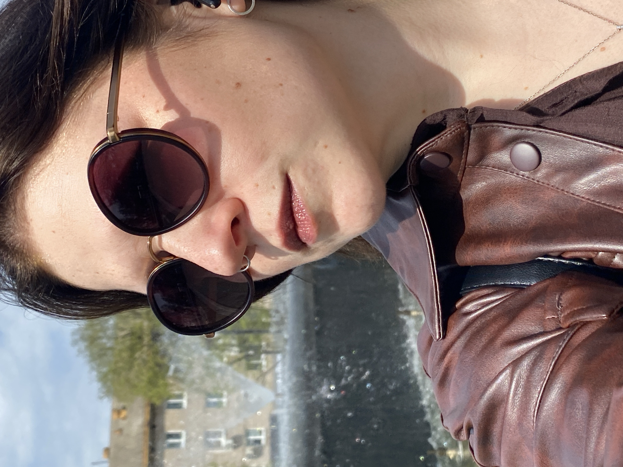 A person is wearing sunglasses and a brown leather jacket. A body of water, likely a fountain, is in the background, along with an apartment building. The image appears to have been taken in the city of Chaikovsky, Russia.