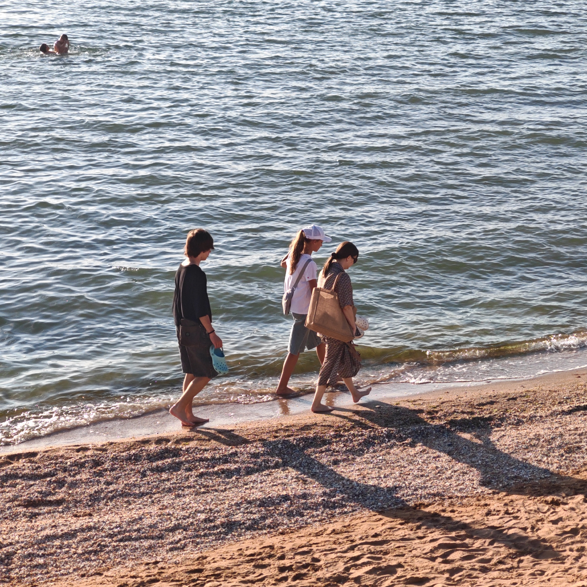 In the city of Shchyolkino, Ukraine, three individuals are walking along a beach next to the sea. A young person with short brown hair, wearing a black t-shirt and shorts, walks on the left, holding a blue sandal. To their right, a person wearing a white cap and a white t-shirt with a grey bag over their shoulder walks beside a third person. This person wears a patterned dress and carries a large beige tote bag, with a white shoe peeking out of it. In the distance, two figures are partially submerged in the water. The sea has small waves and reflects the sunlight. The beach is covered in sand and small shells, with shadows cast across it.
