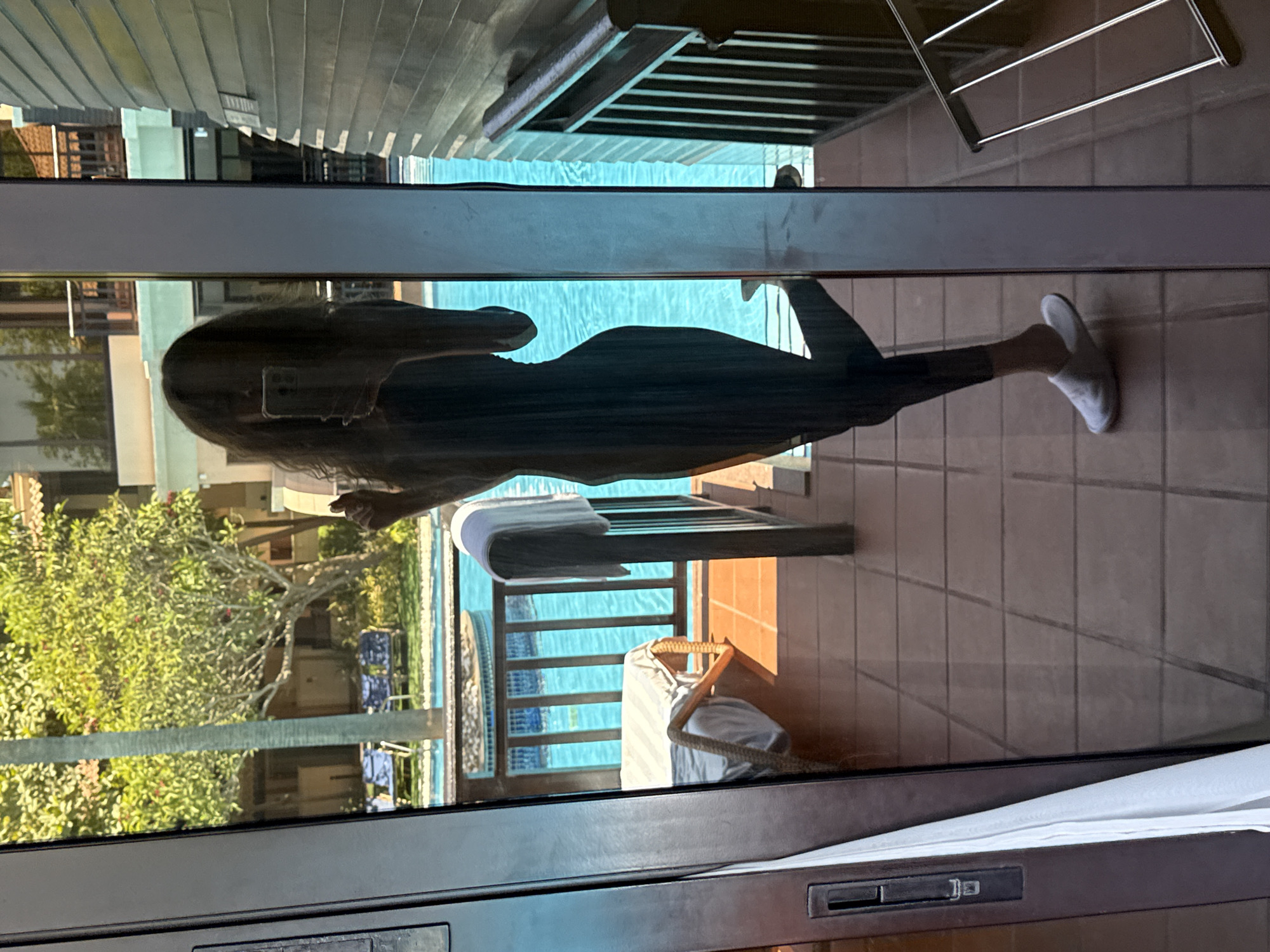 A silhouette of a person is visible in the reflection of a glass door. The person is holding a phone and appears to be taking a selfie. They are wearing dark clothing and white slippers. The reflection shows a poolside area with lounge chairs and palm trees. The environment appears to be a resort or hotel in Dubai, United Arab Emirates. The lighting suggests it is daytime, with sunlight casting shadows on the tiled floor.