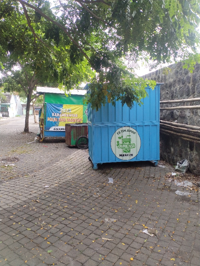 This outdoor scene captures a paved area, likely a street or public pathway, shaded by several green-leafed trees. On the right, a dark stone wall runs along the edge of the paved surface. The ground is made of interlocking square pavers and has scattered dry leaves and some litter, particularly near the wall and the blue stall.
Two portable stalls or carts are prominent. The one closer to the foreground and on the right is bright blue, made of corrugated metal. It has a circular white logo on its side with green text that reads "ES TEH JUMBO", "DIMENSI", and "MURAH CK". Further back and to the left is another stall, green and yellow, with a fabric banner draped across its front. The banner displays the text "2023", "KARANGANYAR", "MAJU DAN MANDIRI", and "DOLANAN". Behind this stall, a smaller, rustic-looking cart with wooden wheels is partially visible.
No people are present in the image. The time of day appears to be sunny daytime, with bright light filtering through the tree canopy, creating dappled shadows on the ground. The location is consistent with a public area in Karanganyar, Indonesia, as indicated by the text on the banner. The presence of the stalls suggests a commercial setting where food or drinks might be sold.