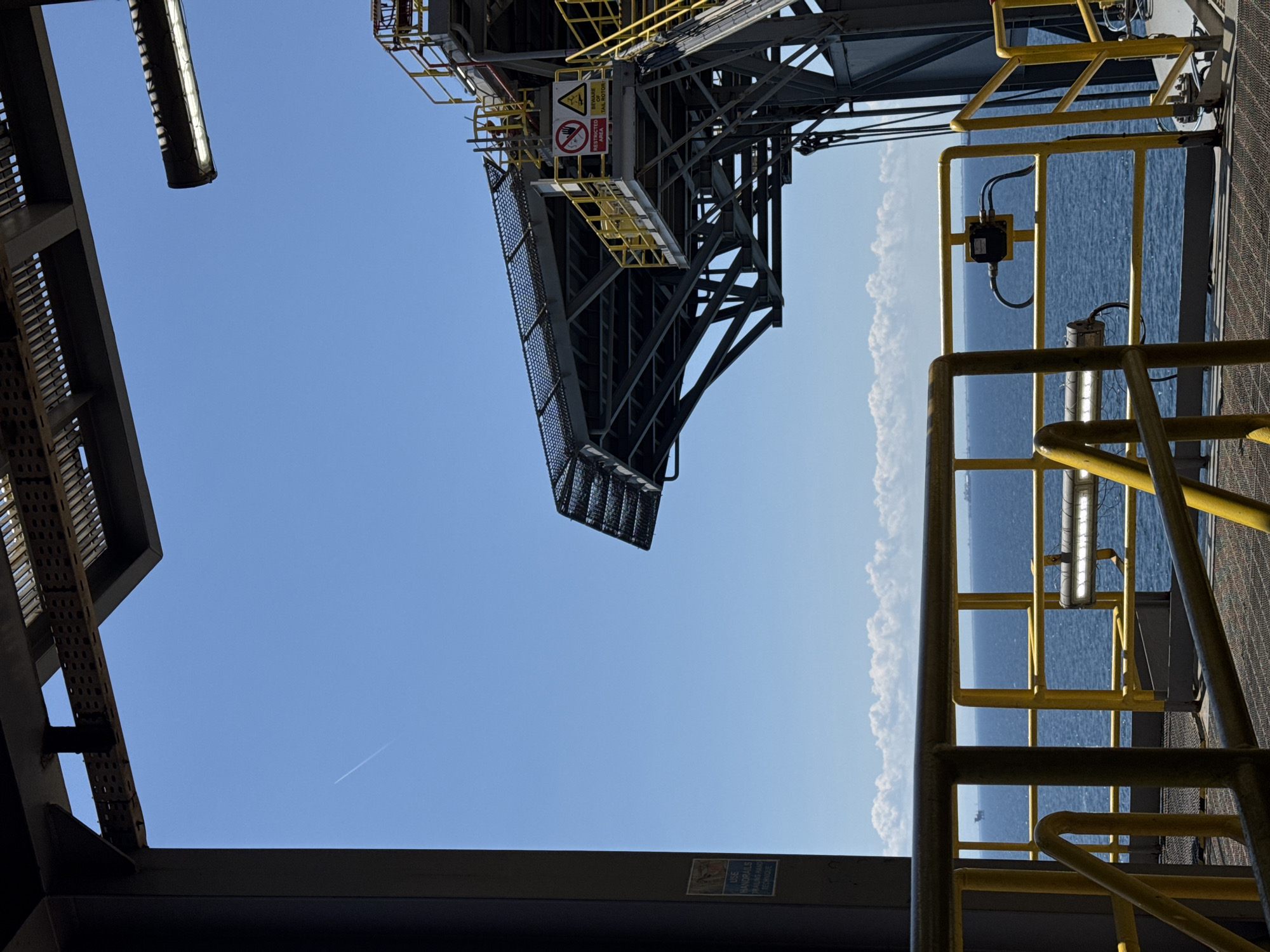 The image depicts an industrial offshore platform under a clear blue sky. In the foreground, yellow metal railings and walkways are visible, suggesting a structured environment. A large metal structure, possibly part of the platform's superstructure, extends from the left side of the frame. In the mid-ground, a helideck is prominent, characterized by its grated surface and supporting metal framework, with yellow safety railings. Two warning signs are affixed to the helideck structure: one shows a red circle with a diagonal line over a hand, indicating "RESTRICTED AREA," and another is a yellow triangle with a black symbol and text that reads "BEWARE TAIL ROTOR." The background reveals a vast expanse of blue ocean, dotted with several distant ships, and a line of white clouds on the horizon. The presence of a contrail from an aircraft is visible high in the sky. The scene is devoid of any people. The overall impression is one of a functional, operational industrial site situated in a marine environment.