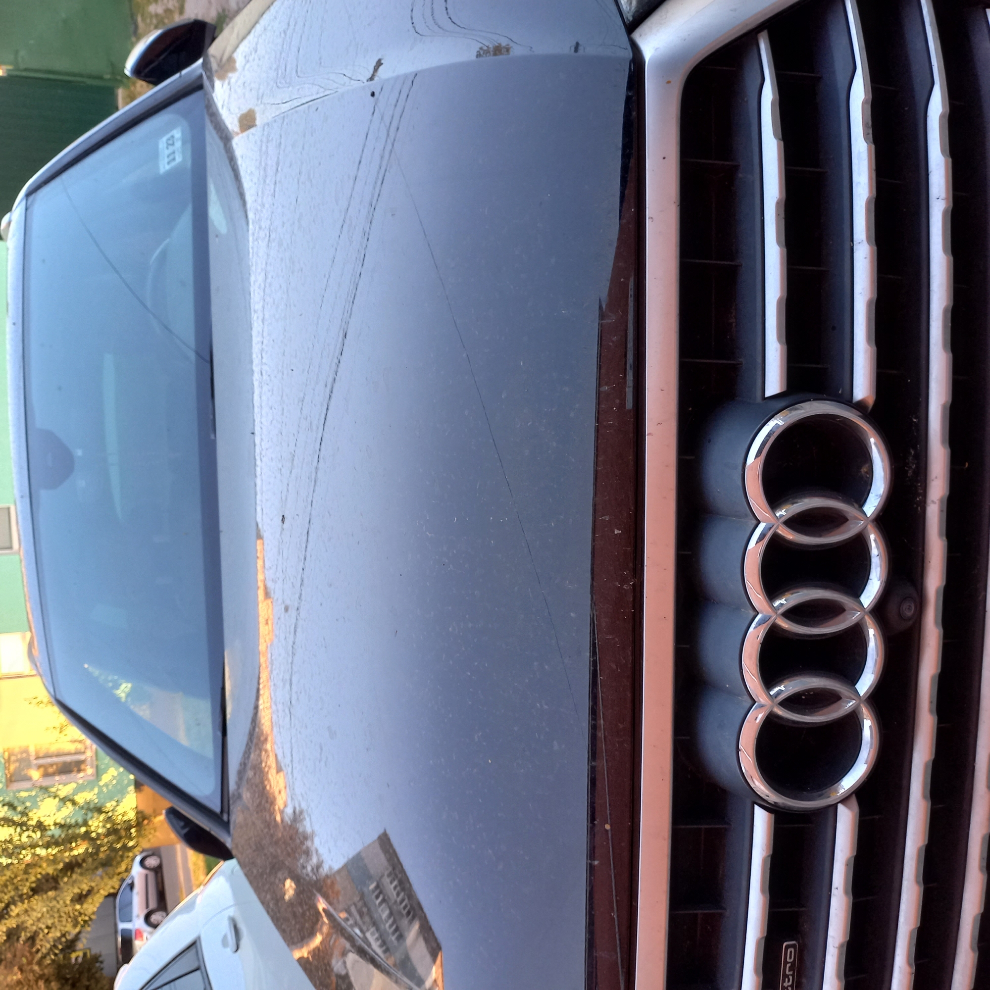 A black Audi SUV is parked on a street in Kropyvnytskyi, Ukraine. The front grille, featuring the signature four rings logo and "quattro" badging, is prominent in the foreground. The hood of the vehicle is dusty, and a crack is visible on the windshield. A building with light-colored walls and a green roof is visible in the background, along with a tree with yellow leaves. Another car, a silver SUV, is parked further down the street. The lighting suggests it is daytime, with sunlight illuminating the scene. There are no people or animals visible in the image.
