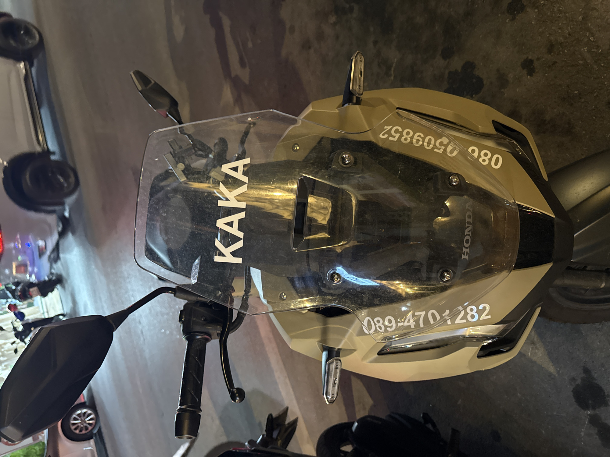The image shows the front of a tan and black motorcycle parked on an asphalt surface, likely at night as indicated by the artificial lighting casting shadows. The motorcycle has a clear windshield with the word "KAKA" written in white, capitalized letters. Below the windshield, a "HONDA" logo is visible. On the lower part of the fairing, two phone numbers are displayed in white lettering: "089-4701282" on the left side and "086 0509852" on the right. The motorcycle is oriented facing forward. In the background, parts of other vehicles, including a white SUV and other motorcycles, are visible, suggesting a public area such as a street or parking lot. The overall scene is that of a parked motorcycle in what appears to be a city environment.