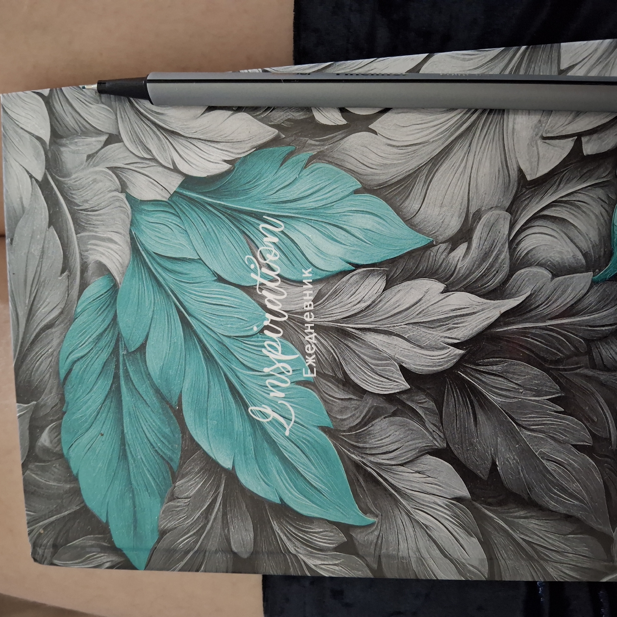 An overhead shot shows a grey and teal journal with a floral leaf pattern. The word "Inspiration" is written in white script across the teal leaves, with the word "Ежедневник" (which translates to "Diary" or "Notebook" in Russian) below it. A grey marker pen with a black tip is positioned vertically on the right side of the journal, obscuring a portion of the design. The journal is resting on a dark, textured surface. The background is a muted beige. The location of this scene is unknown.