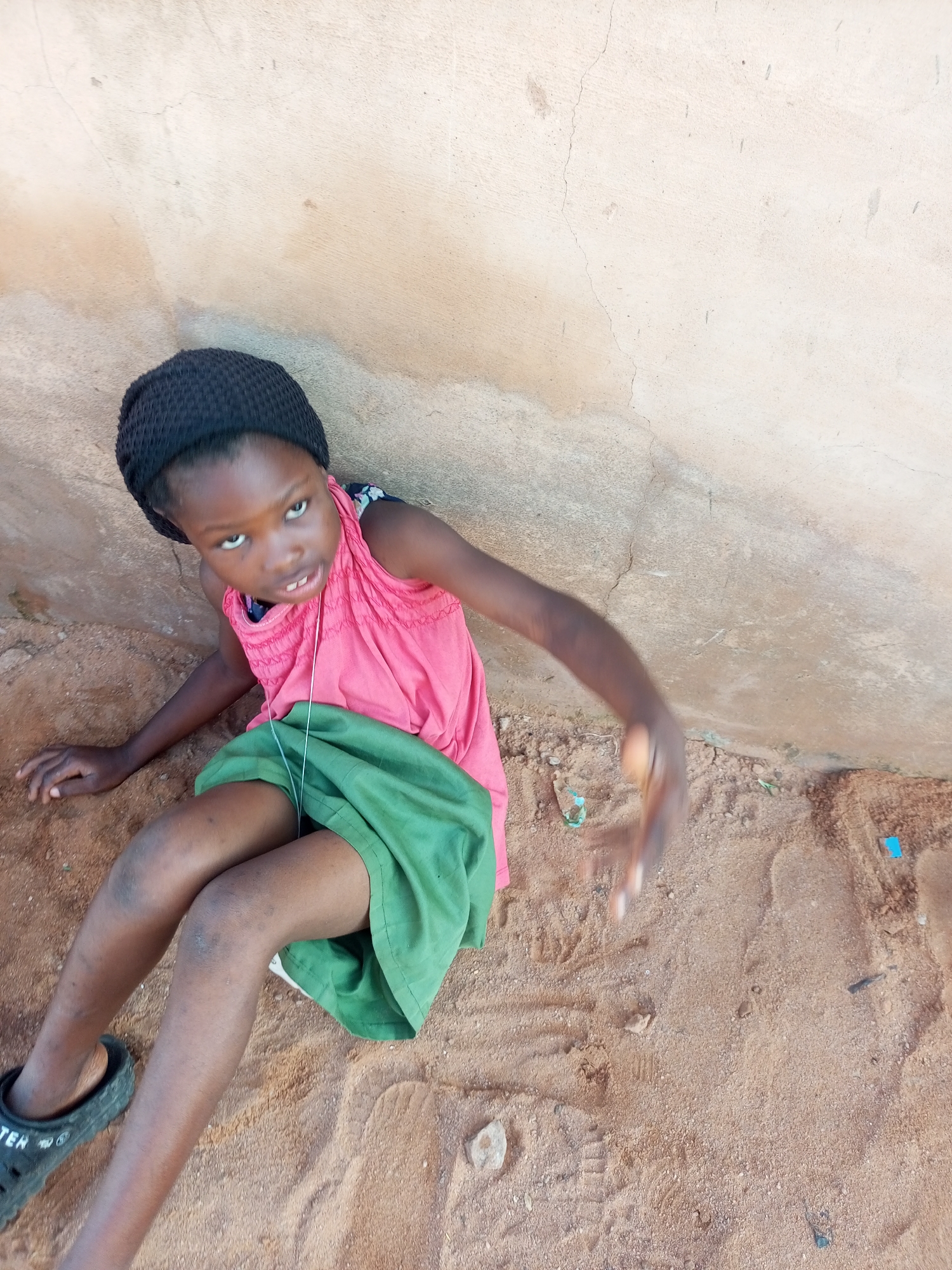 A young girl wearing a black beanie, a pink sleeveless top, and a green skirt is sitting on the sand in front of a stucco wall. Her left hand is resting on the ground, and her right arm is extended forward, with her hand blurred due to motion. She is wearing a black sandal on her left foot. The wall behind her is textured with cracks and discolored patches. The ground is sandy and contains small stones and a few pieces of debris, including a blue plastic object. The scene appears to be outdoors, possibly in a residential area.