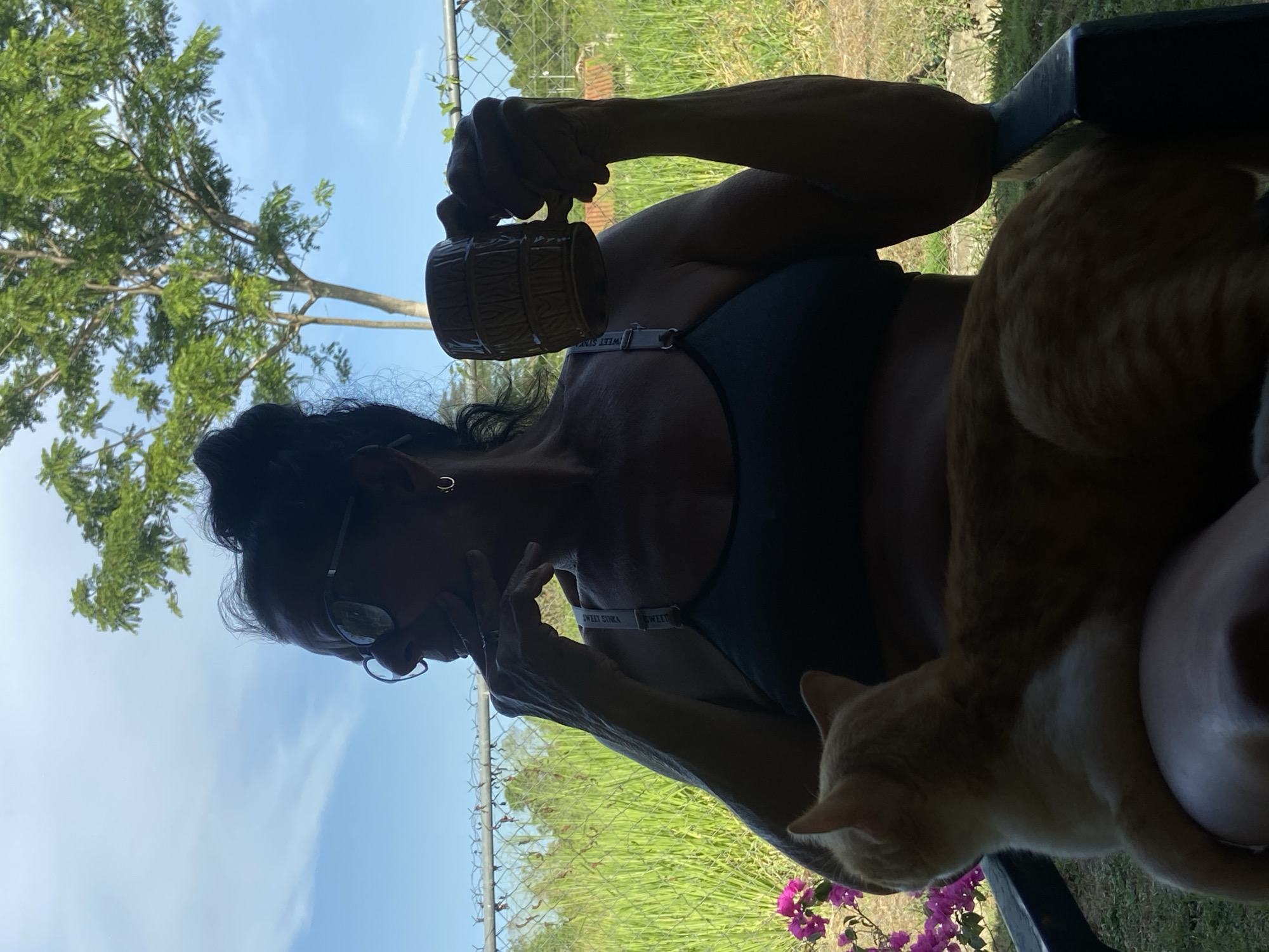 A woman sits outside in an environment with green vegetation and a tree visible, possibly in a yard or garden. She is holding a mug and appears to be either thinking or on the phone. An orange cat is sitting on her lap. The scene is outdoors, and the presence of a chain-link fence suggests a residential or property boundary. Los Velásquez, Venezuela is not explicitly verified in the image but is asserted as the location.