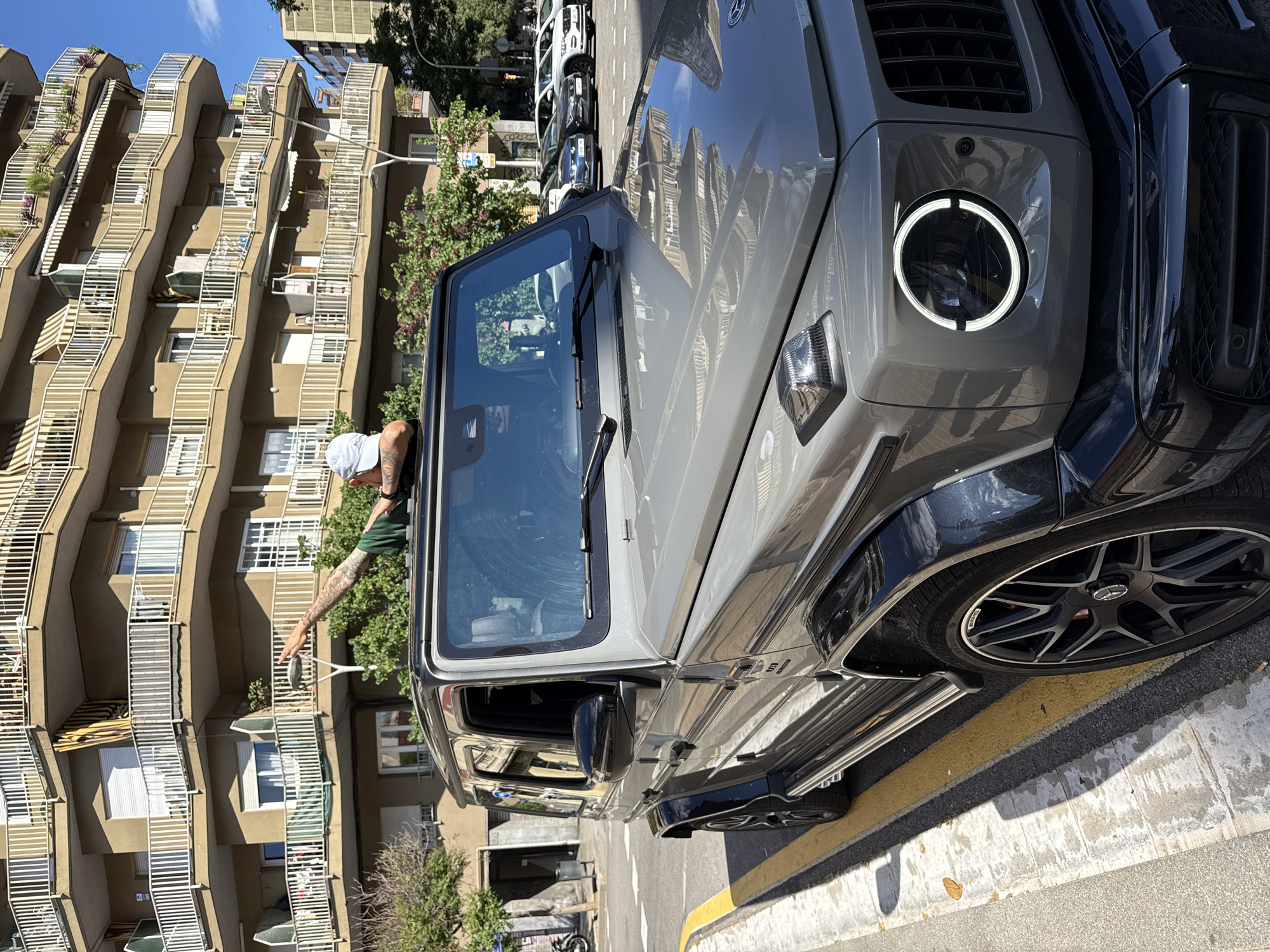 A gray Mercedes-Benz G-Class is parked on a city street, with a man leaning out of the driver's side window. The man is wearing a light-colored baseball cap, and a green shirt with tattoos on his arm. He appears to be interacting with something outside the car, possibly someone or something on the street. The vehicle is parked near a sidewalk with a yellow painted line. In the background, there is an apartment building with unique architectural features and several balconies. Additional vehicles are parked further down the street. The picture was taken in Barcelona, Spain.