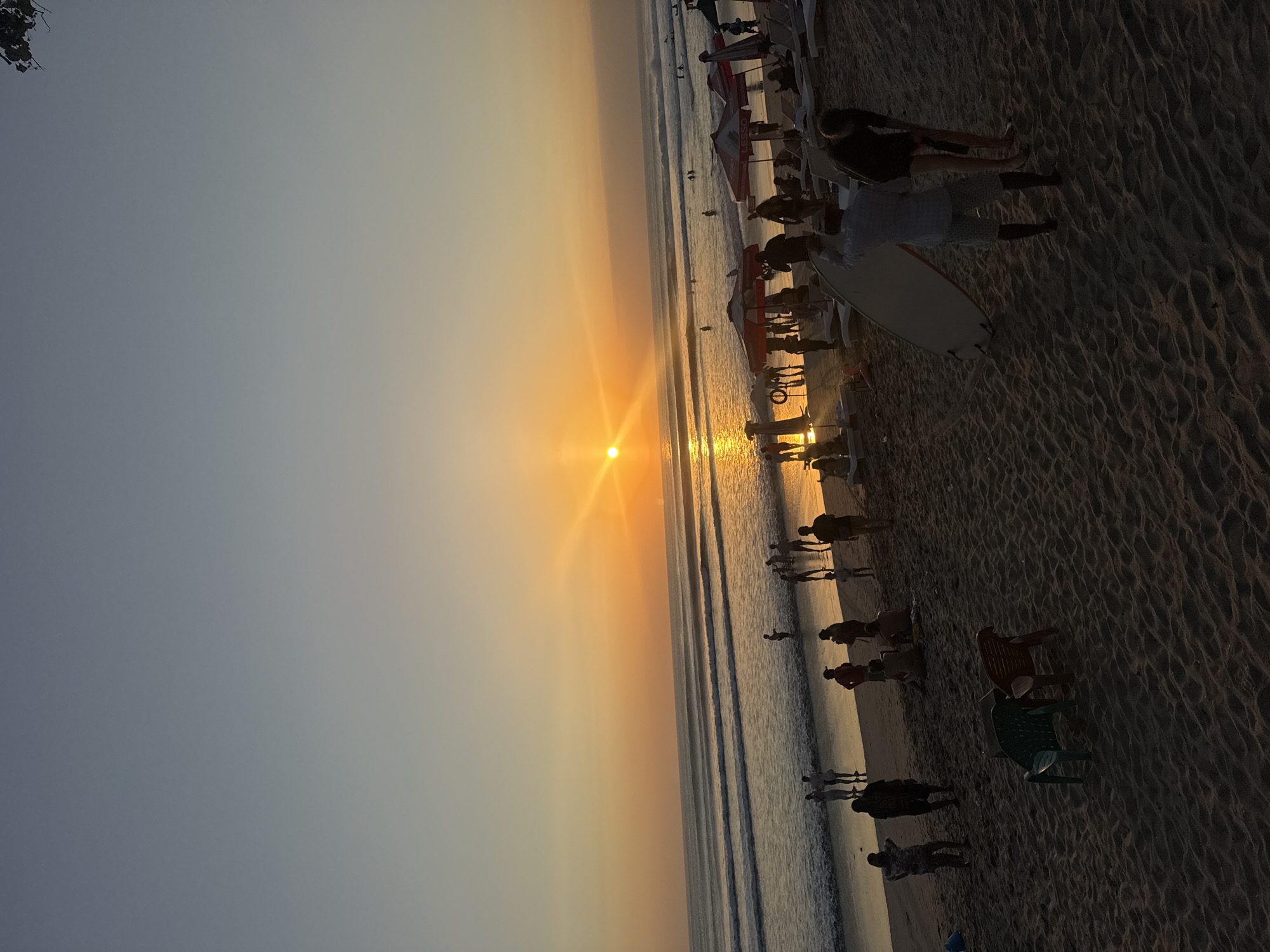 A wide-angle, eye-level shot captures a sunset scene on Kuta Beach, Indonesia. The sky transitions from a pale blue at the top to a vibrant orange near the horizon, where the sun is prominently visible. The sun's rays fan out across the sky and reflect on the calm ocean waves.
The beach is populated with numerous individuals and various objects. In the foreground, on the sandy beach, are two empty beach chairs, one green and one red, positioned close together. Further back, several people are seen walking and interacting. A person in the mid-ground is carrying a surfboard. There are also numerous beach umbrellas and lounge chairs set up.
In the water, several people are swimming or wading, and a few surfers are visible further out. The scene depicts a lively atmosphere with people enjoying the beach at sunset.