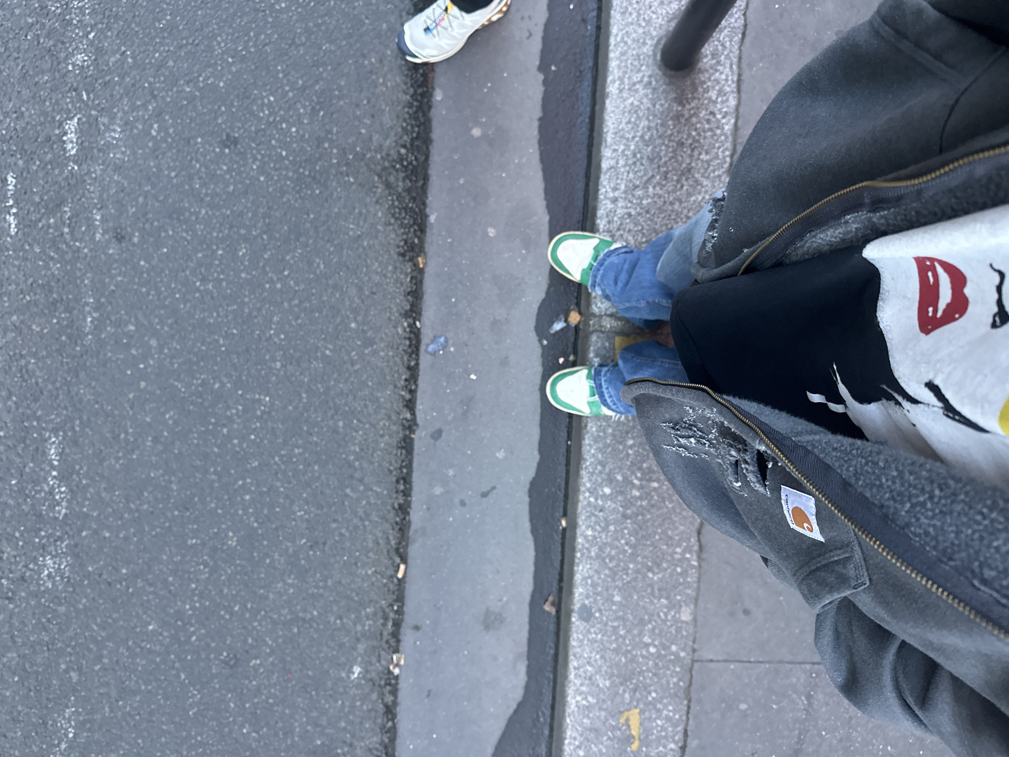 This image, shot from a high angle, shows an urban street scene. The ground is composed of asphalt and concrete. On the right side of the frame, there's a person wearing blue jeans, green and white sneakers, a black shirt, and a gray Carhartt jacket. Another person is visible, wearing white sneakers. There are hints of a metal pole, possibly a bollard or signpost. Given the architecture, paving, and the clothing style, it's likely this image was taken in a European city, possibly Paris, France. There is no other evidence to corroborate this location. The overall environment appears to be a typical urban setting. There are discarded cigarette butts on the ground.