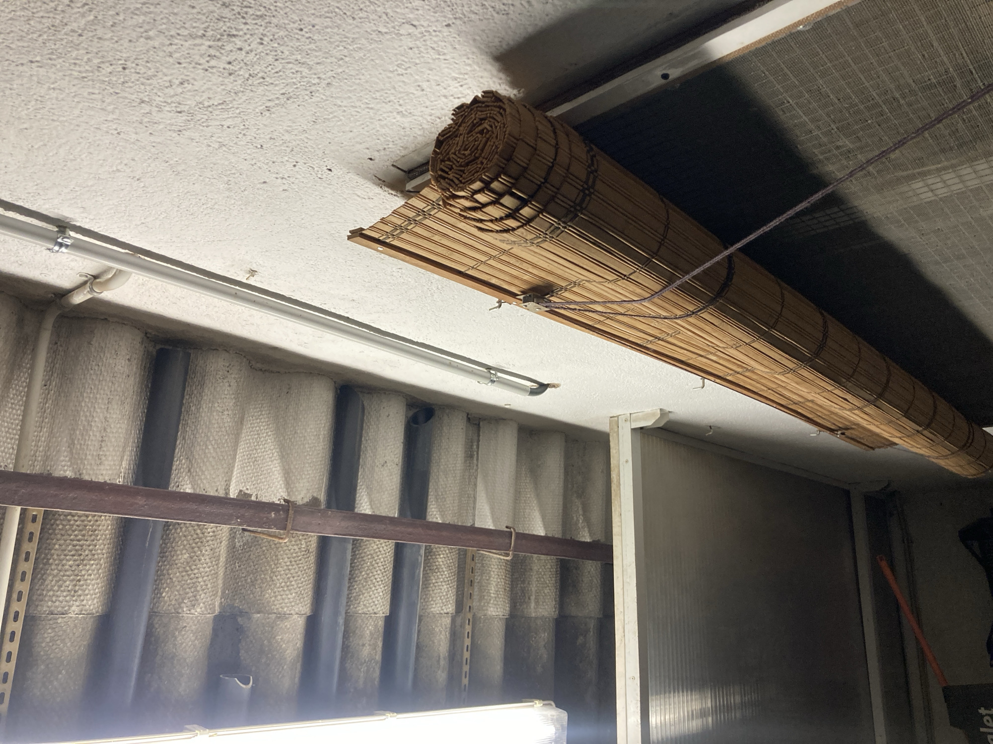 The image shows a close-up view of an interior space, likely a garage or storage area, in Sabadell, Spain. The scene is dominated by a rolled-up bamboo blind mounted on a textured white wall. Above the blind, a corrugated ceiling made of light-colored material is visible, supported by dark horizontal beams and a rusty metal pipe running vertically. To the left, a fluorescent light fixture is partially visible. A metal frame structure, possibly a shelf or partition, extends across the bottom left of the frame. The overall impression is one of a utilitarian, perhaps unfinished, space.