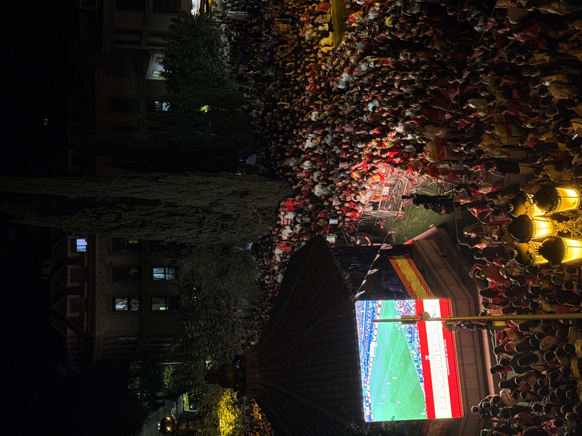 In an outdoor urban setting at night, a large crowd of people, estimated to be in the hundreds, are gathered to watch a soccer match being broadcast on a large LED screen. The screen displays a green soccer field with players in uniform. The crowd is densely packed, with many individuals wearing red and yellow clothing, likely in support of a Spanish team. Some people are holding Spanish flags. The scene is illuminated by the bright screen and ornate streetlights. In the background, there are buildings and trees. The text on the screen, "LAS ROZAS CON GABI" and "Ayuntamiento de Las Rozas" indicates the location as Las Rozas de Madrid, Spain, and suggests a civic event or celebration. A police officer can be seen near a barrier in the foreground.