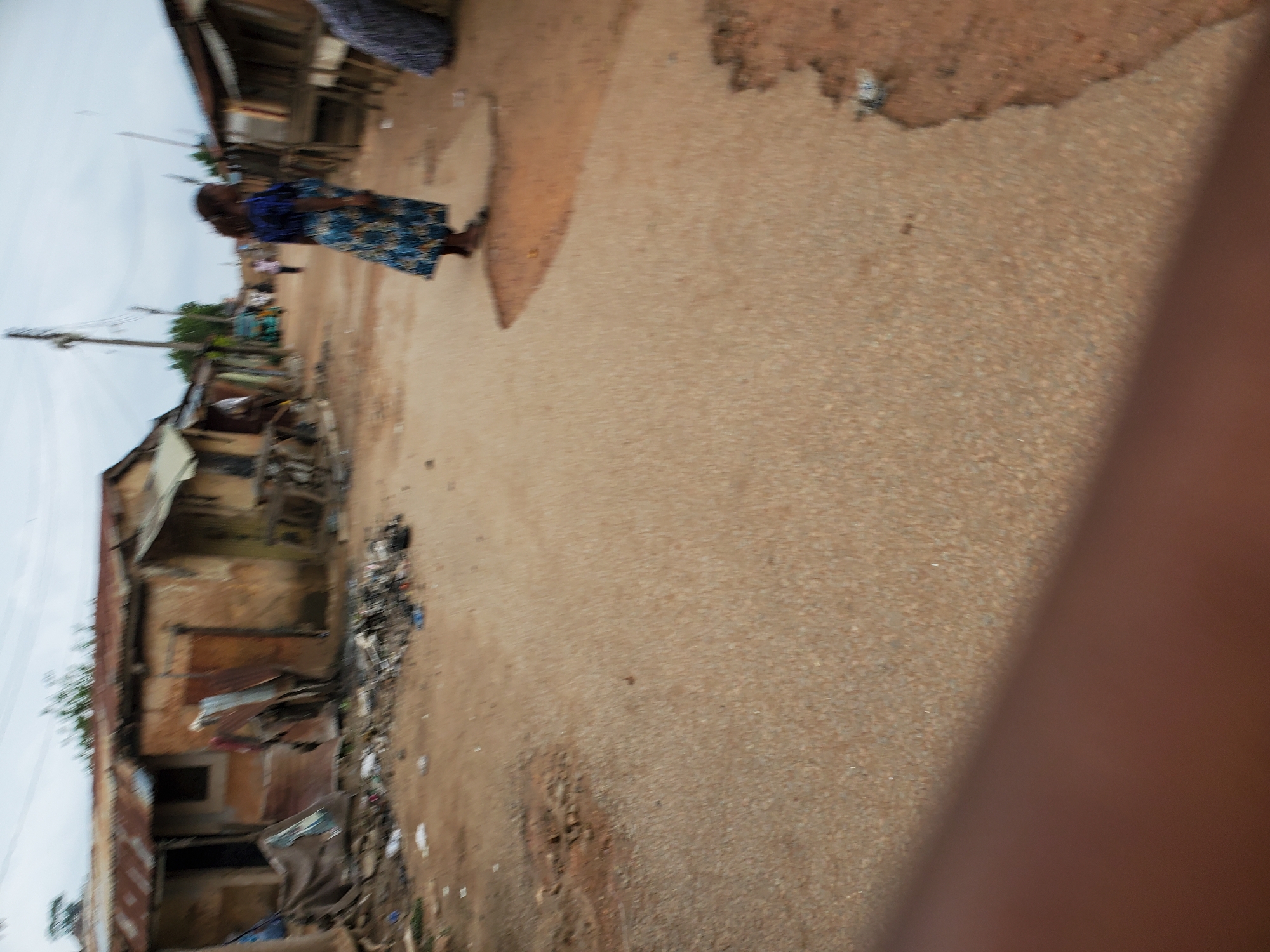A dirt road runs through a slum neighborhood in Jos, Nigeria. Buildings made from corrugated metal and discarded materials line the street, piled with garbage. An African woman in a blue print dress walks down the street. A brown arm is visible in the foreground. Overhead power lines can be seen.