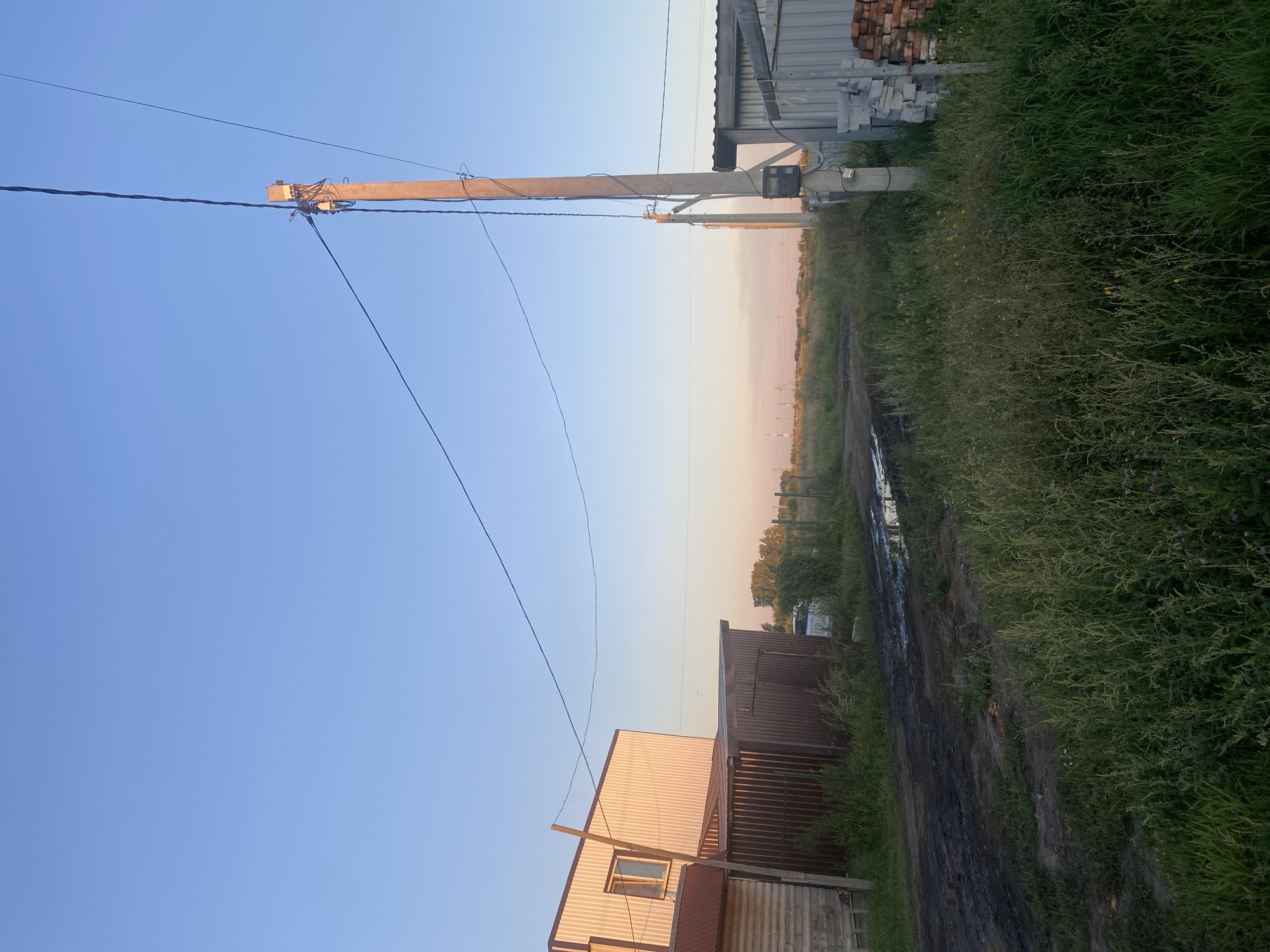 The image depicts an outdoor scene on a dirt road in a rural area, likely on the outskirts of Verkhniy Karbush, Russia, given the context. The sky is clear and pale blue, transitioning to a soft pinkish hue near the horizon, suggesting either sunrise or sunset.
On the left side of the frame, there is a light-colored, corrugated metal building with a red-roofed extension and a wooden section at the base. Adjacent to it is a dark brown corrugated metal shed. The road in front of these structures is muddy and appears to have several puddles, indicating recent rain or poor drainage. Tall, green grass and weeds line the sides of the road.
Dominating the right side of the image is a concrete utility pole supporting electrical wires that stretch across the frame. Attached to the pole is a small electrical meter box. Further to the right, a section of a gray corrugated metal structure is visible, with stacks of light gray building blocks and red bricks next to it. In the background, a white vehicle is partially obscured by greenery. The landscape beyond the road is characterized by more vegetation and a few distant industrial structures or power lines, consistent with a semi-rural or industrial fringe setting. No people are present in the scene.