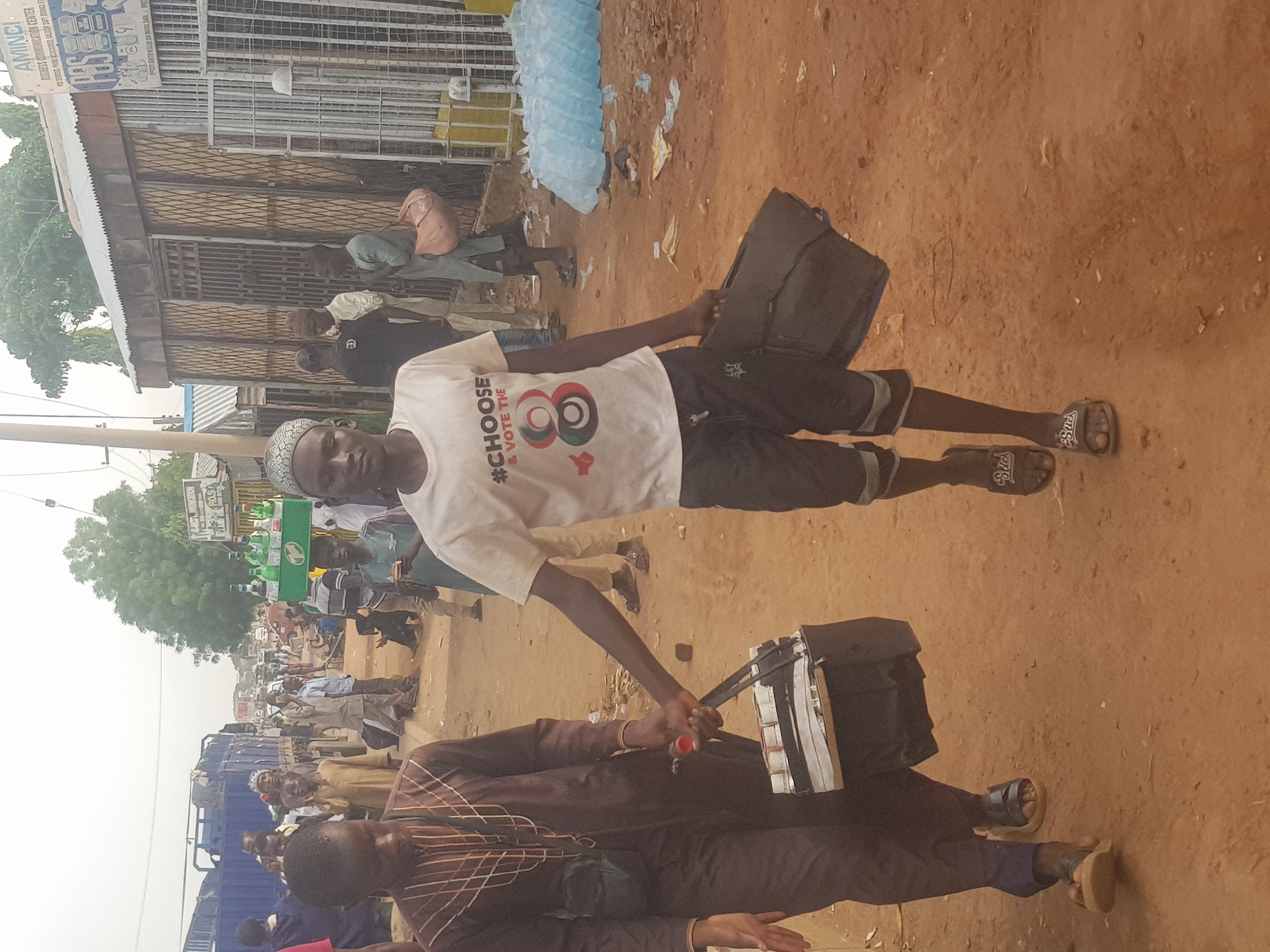 The scene is an outdoor setting, possibly a street or market area in Kufena, Nigeria. The ground is mostly bare earth with a reddish hue. People are scattered throughout the frame, suggesting a public gathering or market. A young man wearing a white t-shirt with political messaging ("#CHOOSE & VOTE THE 8") is holding two black rectangular objects, possibly bags or cases. He is also wearing shorts and sandals. Another man in a brown patterned outfit is in the foreground holding one of the black bags. There is a building with a fenced-in storefront on the left side of the image, and a blue truck in the middle of a crowd. There are trees and general urban clutter scattered throughout the scene.