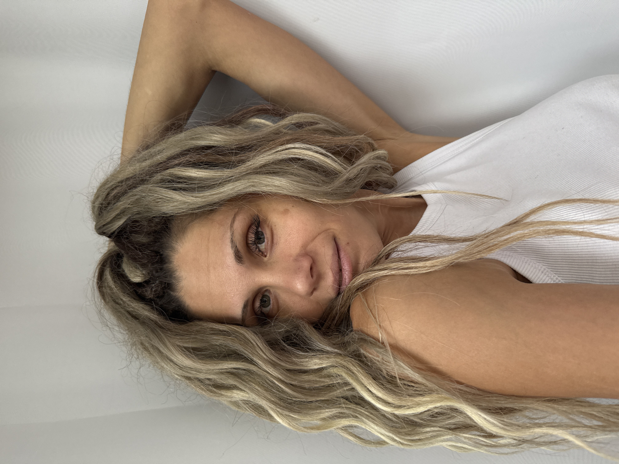 The image shows a woman with blonde, wavy hair pulled up into a high ponytail. She is wearing a white tank top and leaning against a white wall, with her left arm behind her head. Her gaze is directed towards the camera, and she has a slight smile on her lips. The background is a plain white wall, suggesting an indoor setting. There is no indication of the city of Rusanovo, Russia, or any other specific location.