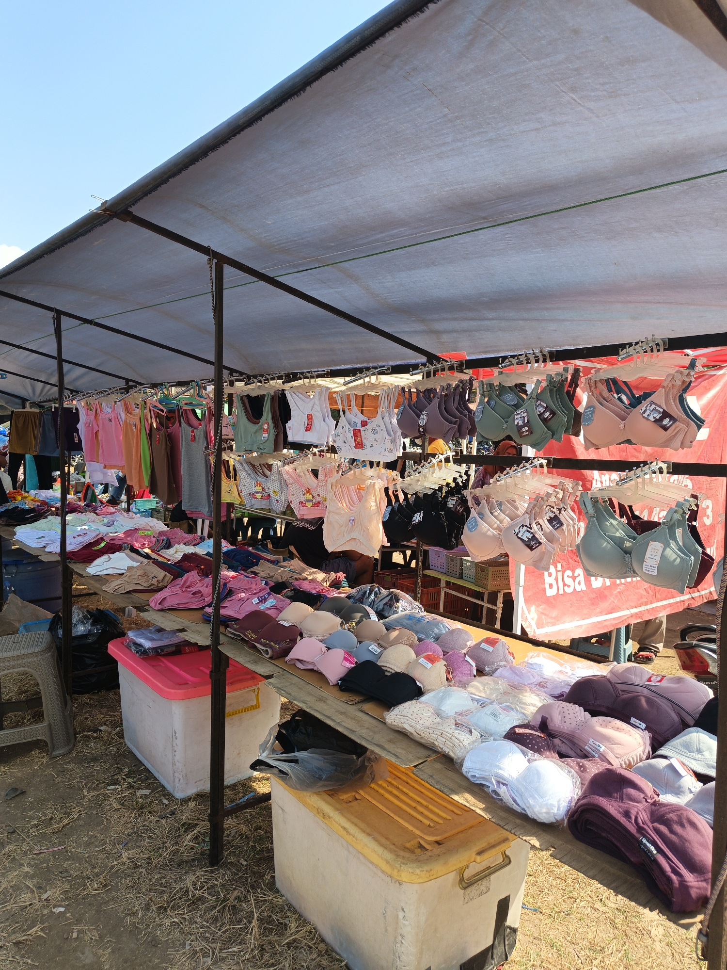 This is a street market scene in Jatinom, Indonesia, under a tarpaulin canopy. The foreground and midground are filled with various clothing items, predominantly bras and tank tops, displayed on tables and hanging from racks. The bras are in a range of colors, including beige, light pink, dusty rose, green, and dark purple. Some are packaged, while others are unboxed and neatly arranged. In the background, more clothing is visible, along with a red banner that has some Indonesian text. The ground is covered with dry grass and dirt, suggesting an outdoor setting. A few plastic containers and a stool are also present. The sky above is clear and blue.