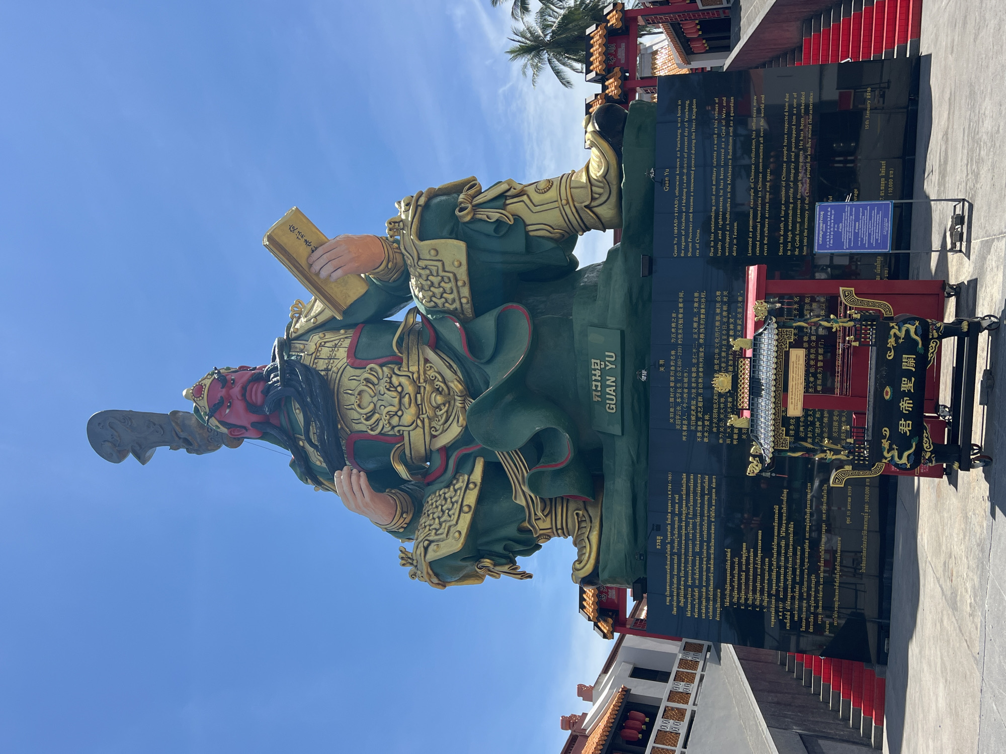 The image shows a large, imposing statue of Guan Yu, a historical Chinese military figure, seated and holding a book. The statue is painted in green and gold, with a red face and a long black beard. It is erected on a black monument with inscriptions in Thai and Chinese. In front of the monument, there is an ornate incense burner, also with Chinese inscriptions. The background features a clear blue sky with some scattered clouds, palm trees, and the facade of a traditional Chinese-style building with red lanterns. The location is identified as the city of Baan Hua Thanon, Thailand, with the statue being a prominent landmark. The scene appears to be an outdoor setting on a sunny day, possibly a temple or a cultural site. There are no people directly visible in the frame interacting with the statue or the monument.