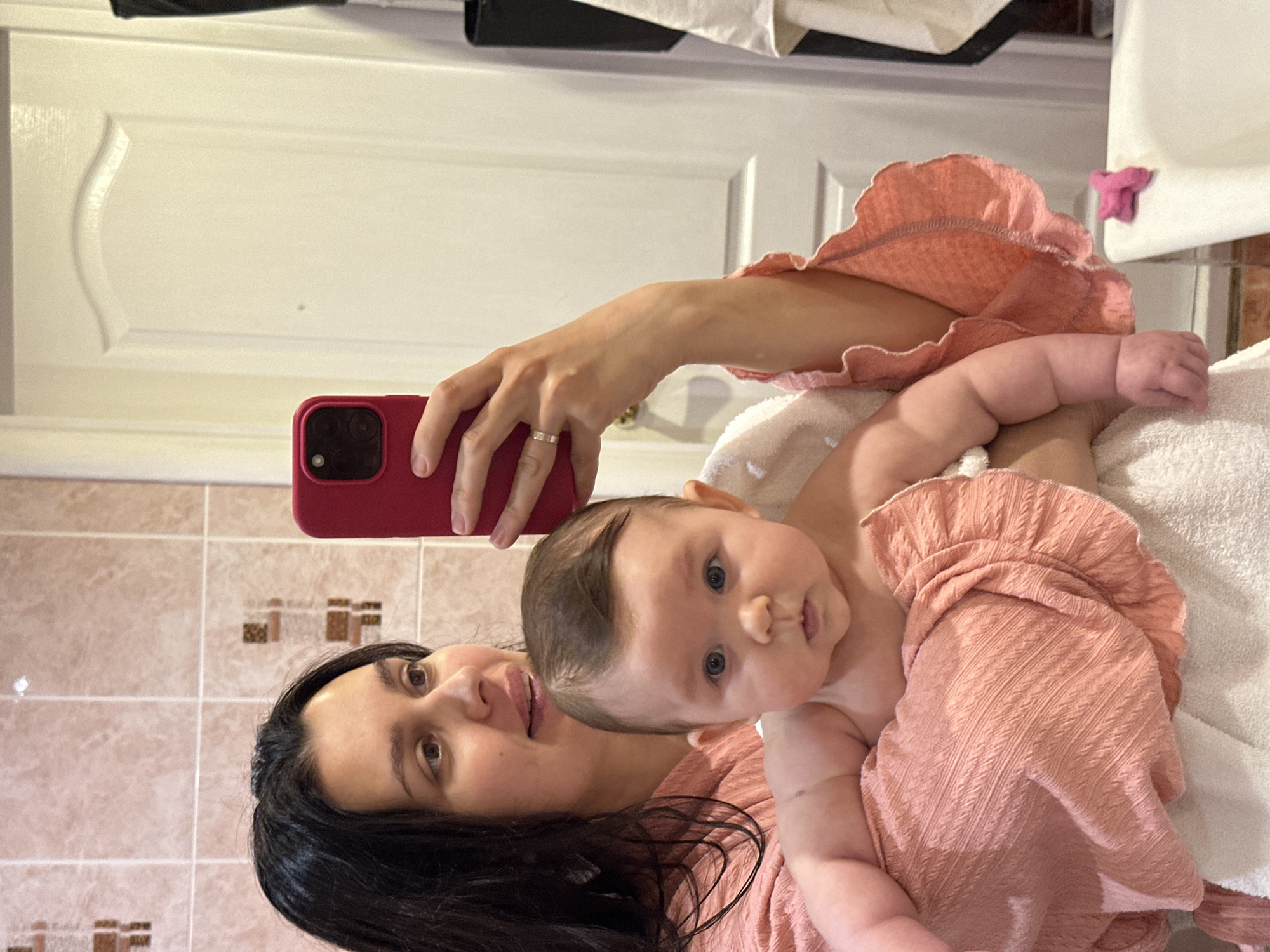 The image captures a mirror selfie taken by a woman holding a baby in what appears to be a bathroom. The scene is oriented sideways, showing the reflection of the subjects.
The primary subjects are a woman and a baby. The woman has long, dark hair and is looking at the camera with a soft, slightly smiling expression. She is wearing a peach or light pink textured top. On her left ring finger, a silver-toned ring is visible. She holds a red smartphone, equipped with multiple camera lenses, which is being used to take the selfie.
Cradled in the woman's arms is a baby, seemingly wrapped in a matching peach or pink textured fabric. The baby has light-colored hair, possibly brown, and prominent blue eyes, looking with a calm and curious gaze directly towards the phone. The baby's chubby arms and small hands are visible above the fabric.
The background elements suggest an indoor setting, specifically a bathroom. Behind the woman, there is a wall covered with light brown square tiles, featuring scattered decorative dark brown tile accents. To the right, a white panel door with a subtle curved molding is visible. A white towel or blanket is partially seen, supporting the baby. Further right, a small, crinkled pink fabric item hangs from a white surface, possibly a sink or vanity.
The activity depicted is a mother taking a mirror selfie with her child, capturing a tender and domestic moment. The emotions conveyed are warmth and affection. There is no visible text in the image. The location context for this scene is Chaikovsky, Russia.