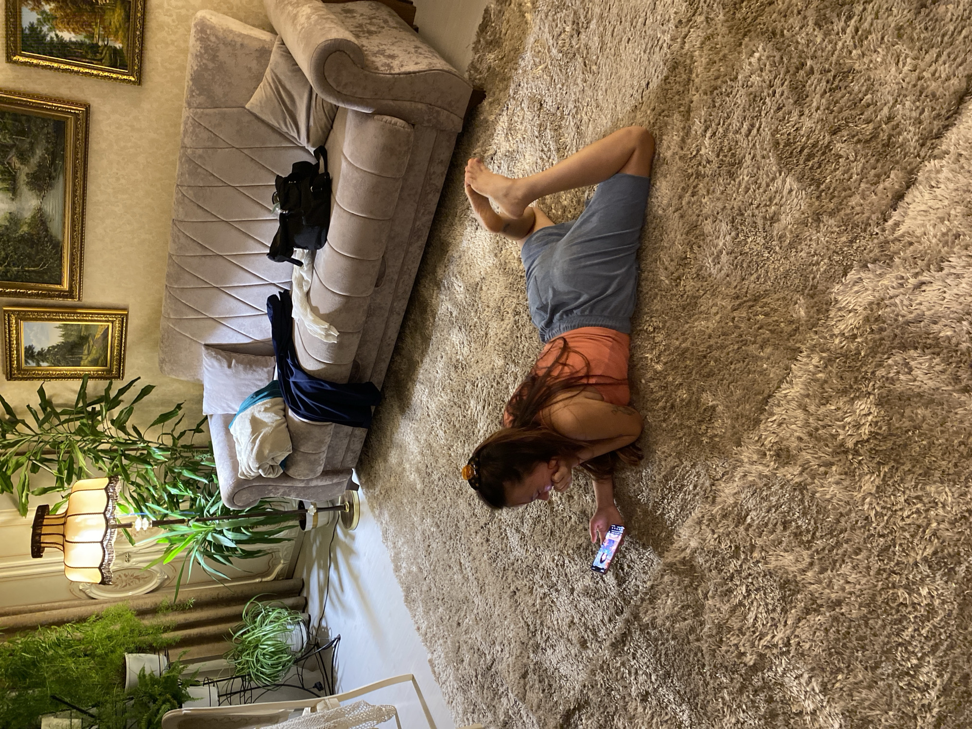 A young woman with long brown hair is lying on a fluffy brown carpet in a room, looking at her smartphone. She is wearing an orange tank top and blue shorts. Her left elbow is bent, and her hand is propping up her chin as she gazes at the screen. Her legs are bent at the knees, with her feet pointing upwards. In the background, a plush light-colored sofa is visible, with folded clothes and a black bag resting on it. Several houseplants are placed around the room, and a vintage lamp with a fringed shade stands near the sofa. The walls are decorated with framed landscape paintings. The scene appears to be an indoor residential setting in Votkinsk, Russia.
