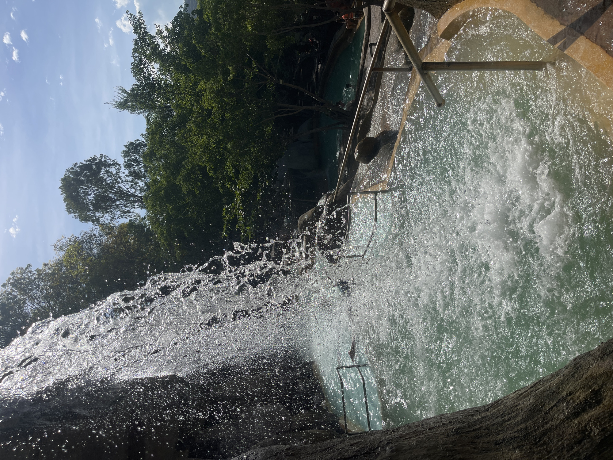 The image depicts a cascading waterfall into a man-made pool. Sunlight glints off the water, creating a shimmering effect. The pool is turquoise and appears to be part of a resort or spa. Several people are visible in the water, some swimming, others relaxing. Railings are installed along the edge of the pool, presumably for safety. Lush green trees and vegetation surround the area, and the sky above is blue with scattered white clouds. The overall impression is one of a serene and inviting outdoor water attraction.