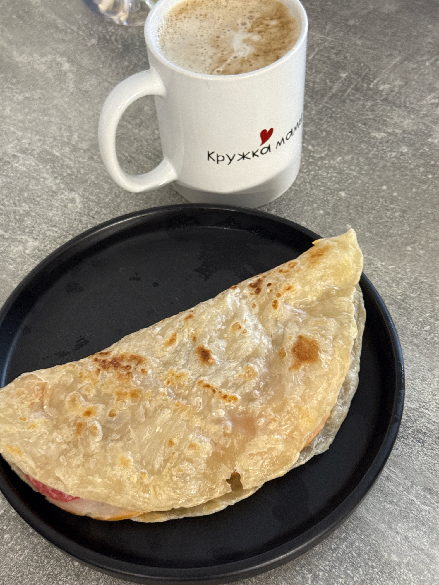 A close-up, overhead shot shows a white mug with a red heart and Cyrillic text, containing a frothy beverage, placed on a gray, speckled countertop. To the left and slightly in front of the mug, a dark, circular plate holds a folded flatbread, possibly a quesadilla or wrap, with visible red and white filling. The lighting is natural and bright, casting subtle shadows. The image is a still life arrangement, likely a breakfast or snack setting. The location is a kitchen or dining area in Lyubertsy, Russia.