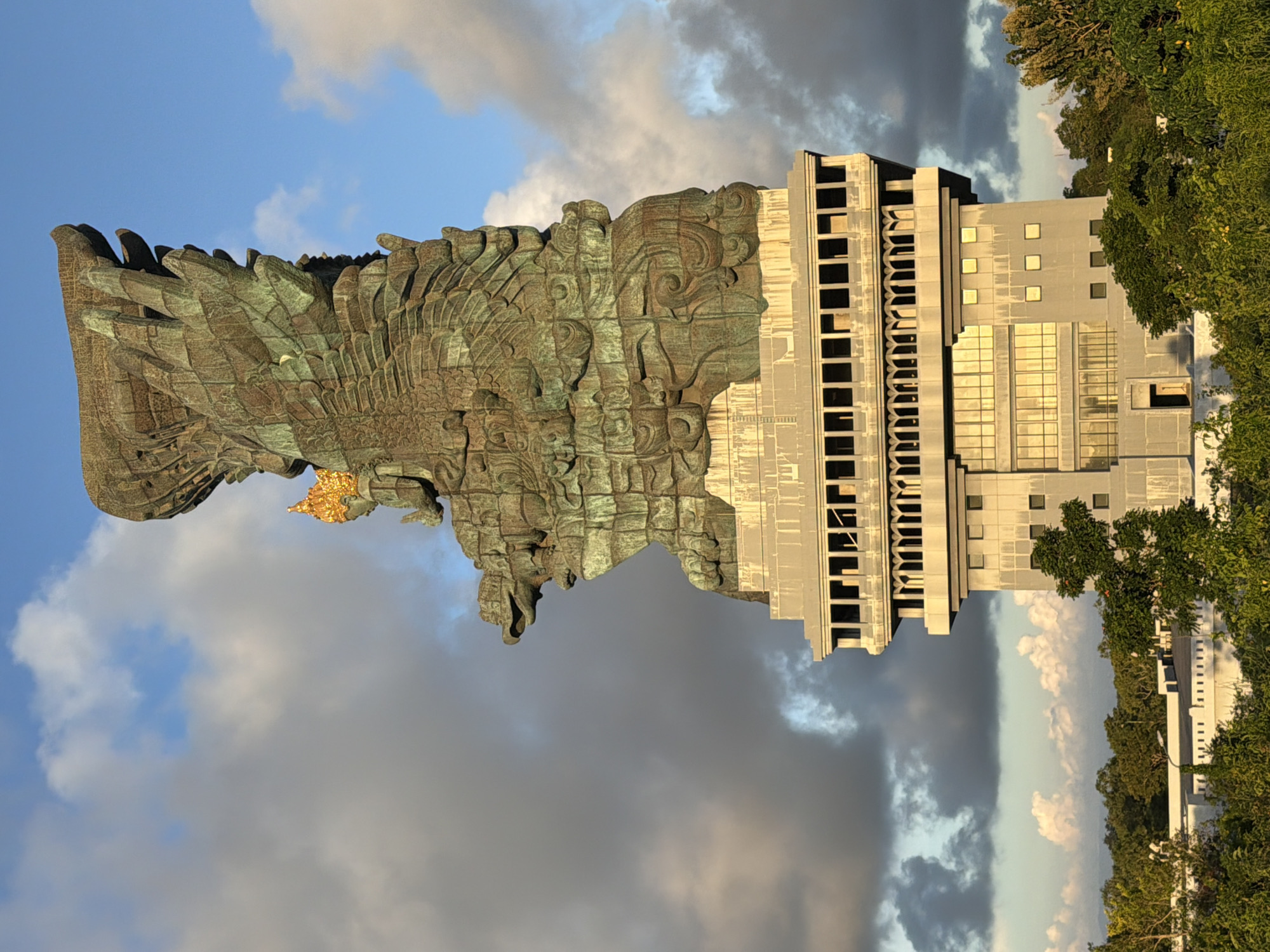 The image depicts the Garuda Wisnu Kencana statue complex in Ungasan, Indonesia, set against a cloudy sky. The statue of Garuda is on the left, rendered in a green-tinged metal. Adjacent to it is the concrete building-like structure of the statue. The right side of the image shows trees and foliage.