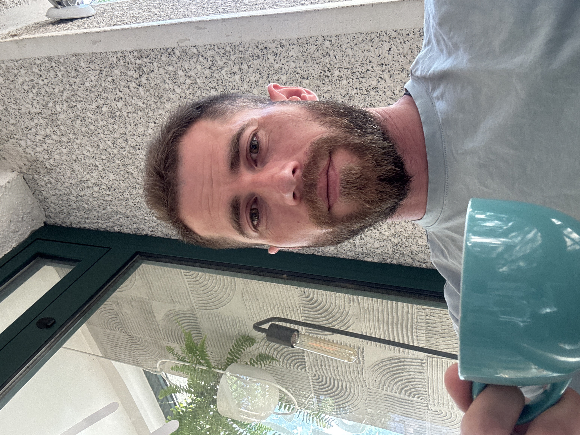 A man with a beard and short brown hair holds a teal mug in his left hand. He is wearing a light blue t-shirt. He is looking at the camera. The background consists of a wall with a textured pattern and a window with a green frame. A plant and a lamp with a visible Edison bulb are also in the background, suggesting an outdoor or semi-outdoor setting. The overall lighting suggests daylight. The scene appears to be from a casual setting, possibly a balcony or patio. The image does not contain any elements that definitively place it in Budva, Montenegro.