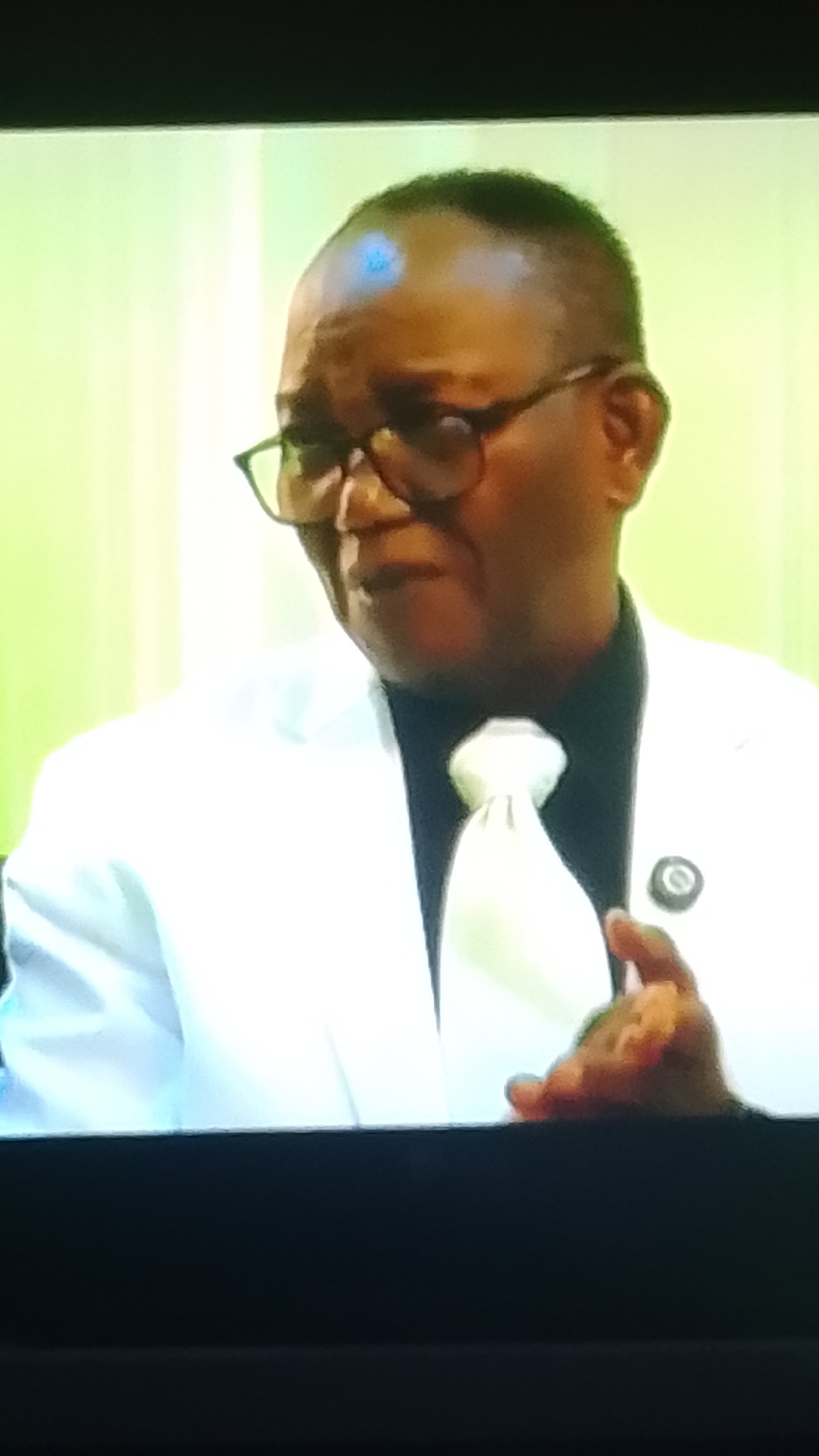 The image shows a medium shot of a Black man wearing a white suit, a black shirt, and a light-colored tie. He is also wearing glasses. The man appears to be speaking or in mid-conversation, with his mouth slightly open and his right hand raised in front of him, gesturing. A round pin is visible on the lapel of his suit jacket. The background is a soft green, possibly a wall or curtain, with vertical lines suggesting a textured surface or lighting. The image is a photograph of a screen, as indicated by the black border at the top and bottom. The location is unknown, as the context of the image does not provide any geographical information.