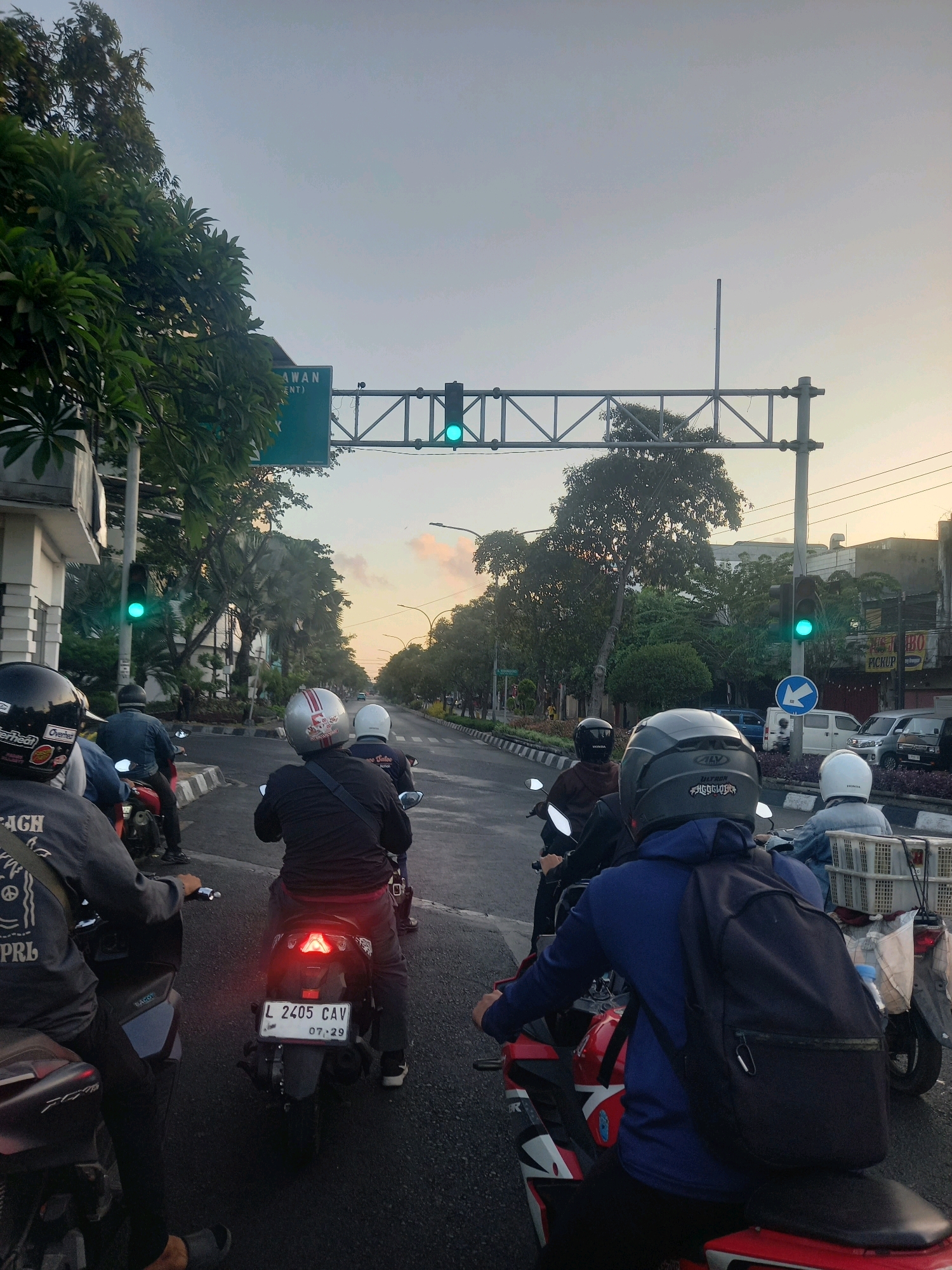 In Surabaya, Indonesia, at dusk, a group of motorcyclists are stopped at a traffic light. The traffic signal displays a green light, indicating it is safe to proceed. The motorcyclists are wearing helmets, with some wearing casual clothing and others wearing more protective gear. The vehicles are a mix of scooters and motorcycles. The road is paved, and the surrounding environment features trees, buildings, and overhead traffic infrastructure. A street sign is visible, with the text "JAWAN" and "(ENT)" on it. The sky shows a gradient of colors, from light blue to hints of orange and pink, indicating the setting sun. The overall atmosphere suggests a typical urban traffic scene at the end of the day.