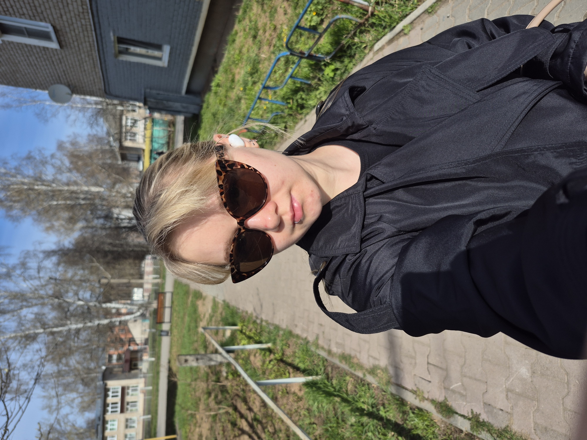 The image shows a person outdoors, likely in a city setting. The person is wearing sunglasses and a black jacket, and has a blonde hairstyle. In the background, there are buildings, trees, and a walkway. The location is likely Chaikovsky, Russia, based on image information.