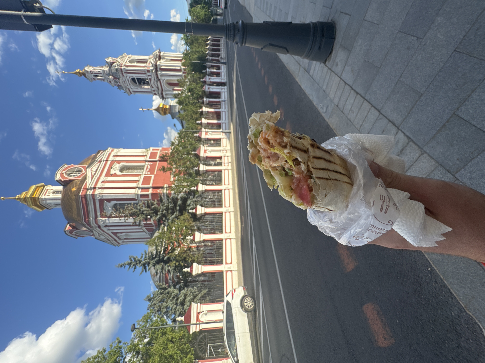 A first-person perspective shot captures a person holding a shawarma wrapped in plastic and a napkin, with a portion of the shawarma and the napkin visible in the foreground. Behind the hand holding the food, a street in Moscow, Russia, unfolds under a bright blue sky dotted with white clouds. The street is paved with asphalt and lined with a sidewalk made of grey cobblestones. A black lamppost stands on the sidewalk to the right of the frame. In the background, across the street, is a picturesque view of the Church of the Resurrection of the Word at the Old Valdaysky Courtyard in Moscow, a prominent architectural landmark featuring red and white facades, gilded domes, and a bell tower. A white SUV is seen driving on the street, and trees with green foliage line the perimeter of the church grounds, separated by a red and white ornate fence.