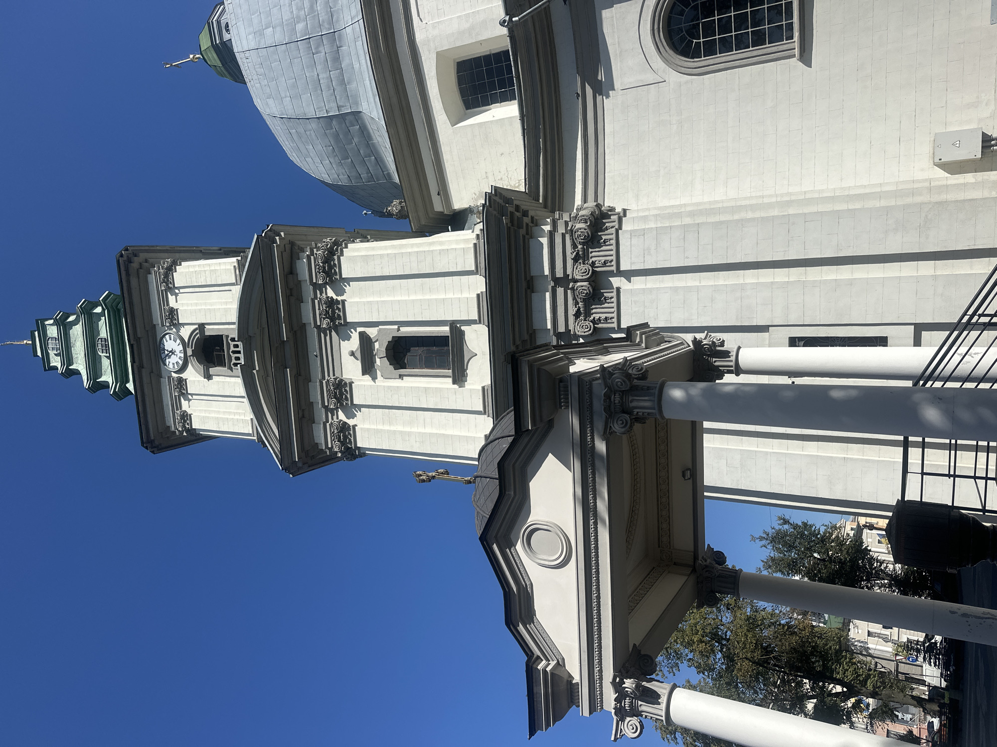 The image shows a low-angle, full exterior shot of the Ternopil Cathedral of the Immaculate Conception of the Blessed Virgin Mary in Ternopil, Ukraine, on a clear, sunny day. The architecture is predominantly white with grey and green accents. A tall bell tower with a clock face and a green spire dominates the left side of the frame. To the right, a large grey dome with a smaller green spire and a cross is visible. The building features ornate classical architectural elements such as columns, cornices, and decorative carvings. A portico with large white columns is in the foreground, leading towards the entrance of the cathedral. In the background, trees and other buildings are visible, suggesting an urban setting. No people are present in the image.