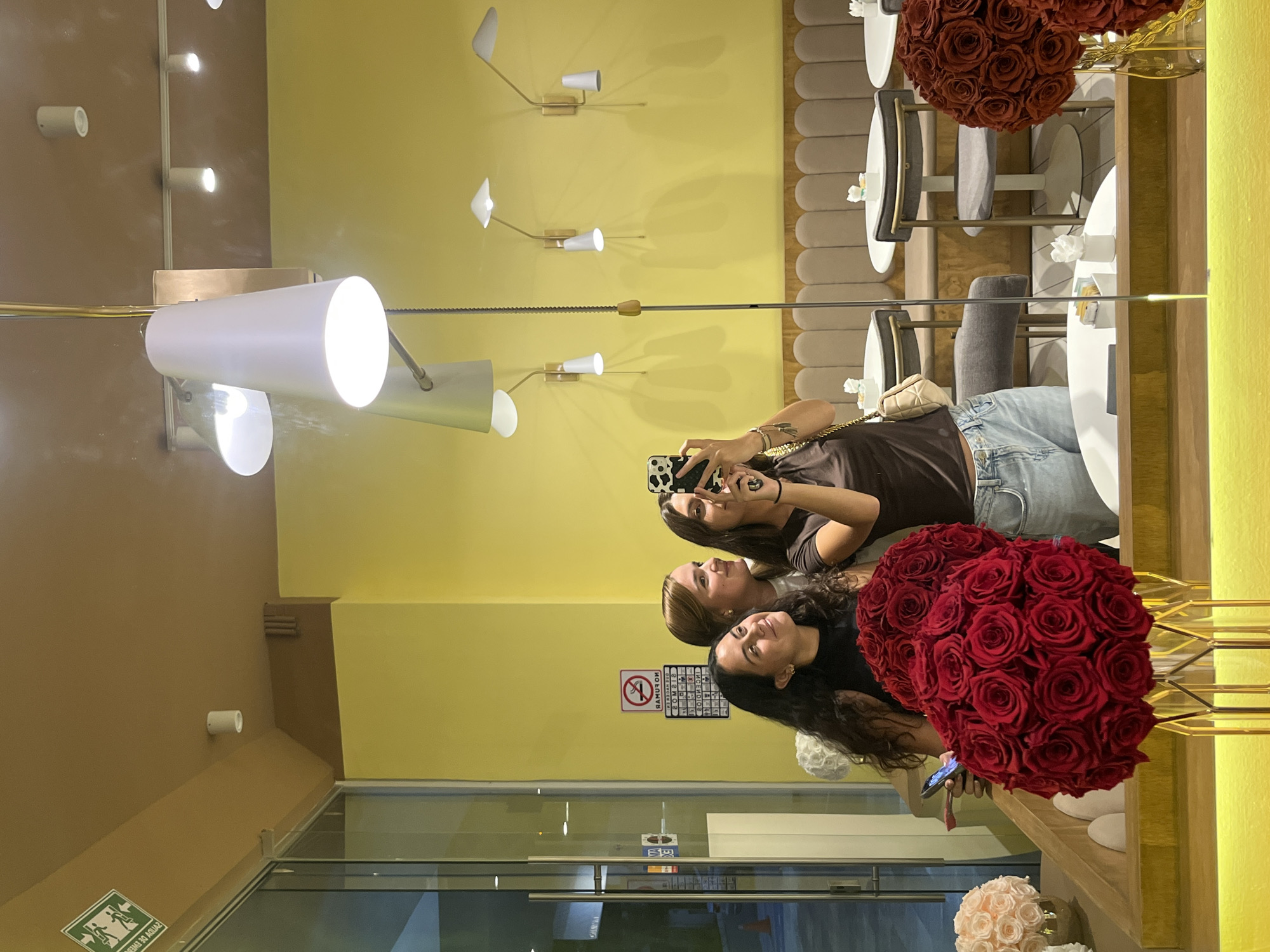 Three women are standing near a reflective surface in what appears to be a retail or dining establishment. The location is most likely in Puebla, Mexico.
A woman in a brown shirt is taking a picture with her cell phone. Behind her, a woman with blonde hair looks up. At the front, a woman with long, dark curly hair smiles.
In the foreground, on the right, there are large bouquets of red roses. In the background, there are tables and chairs. One table appears to have small white flowers on it. The walls are yellow. A white tube-shaped light fixture is present, along with decorative wall lights.
There is a sign with a "no smoking" symbol and some text in Spanish. In the bottom of the frame there is a glass wall.
