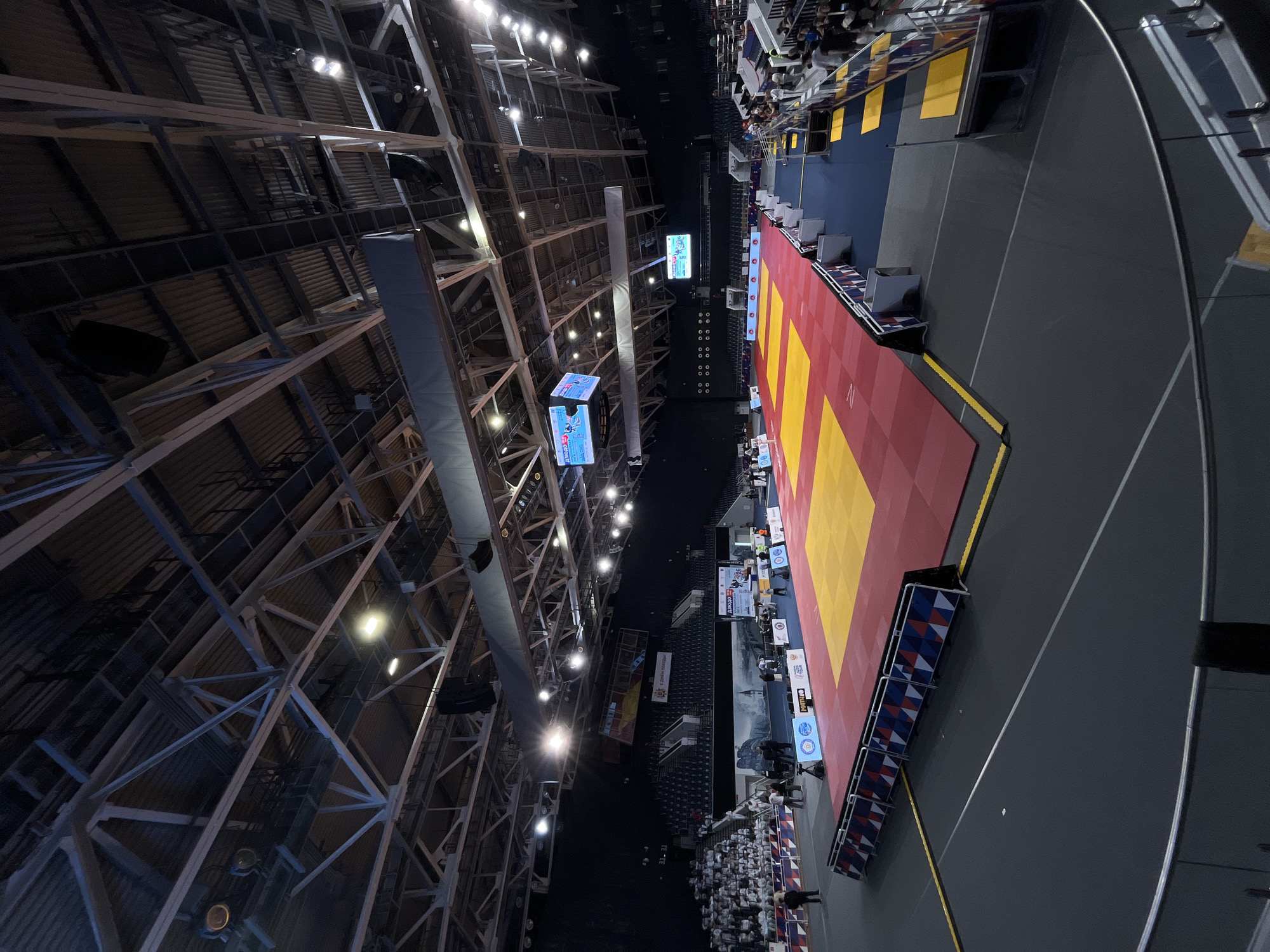 An interior wide-angle photograph captures the Platinum Arena in Krasnoyarsk, Russia, configured for a judo competition. The central focus is an elevated, multi-mat judo area comprised of interconnected red mats with central yellow square sections. This mat area is bordered by low barriers featuring a red, white, and blue geometric pattern. The main floor surrounding the mats is dark grey, marked with white and yellow lines.
Tiered seating, primarily dark blue or black, extends around the arena. While largely unoccupied, a section of seats on the far left contains individuals dressed in white attire, possibly participants or officials. Numerous people, including staff, officials, and photographers, are visible at floor level surrounding the judo mats.
Multiple large LED screens displaying "ДЗЮДО" (Judo in Cyrillic) and other event information are suspended from the ceiling. Additional banners and smaller digital displays bearing Russian text and logos, such as "С Днем Победы," "МАТЧ ТВ," and "RUSSIA," are positioned around the mat perimeter. The arena's high ceiling reveals an extensive network of exposed steel trusses, ductwork, and professional lighting fixtures and speakers. Overhead spotlights illuminate the central competition area, creating strong contrasts with the darker, shadowed upper regions of the venue. The overall scene depicts a professional sporting event setup with limited attendance.