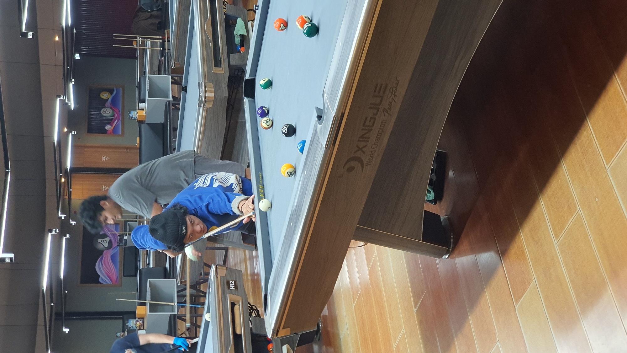 The image shows an indoor billiards hall, likely in Kuta Selatan, Indonesia, with several pool tables. In the foreground, a young person wearing a blue sweatshirt and a black beanie is bent over a pool table, holding a cue stick and aiming to strike a white ball. Their left hand is resting on the table for support. Another person, a young male wearing a gray t-shirt, stands behind and to the right of the person playing, observing the game. Other pool balls are scattered on the table, including a yellow and orange striped ball, a solid orange ball, a solid green ball, a solid purple ball, and a black ball with a white dot. The pool table is a "XINGJUE" brand. In the background, other pool tables are visible, some with people around them. The floor is made of light brown, rectangular tiles. The lighting is provided by fluorescent lights on the ceiling.