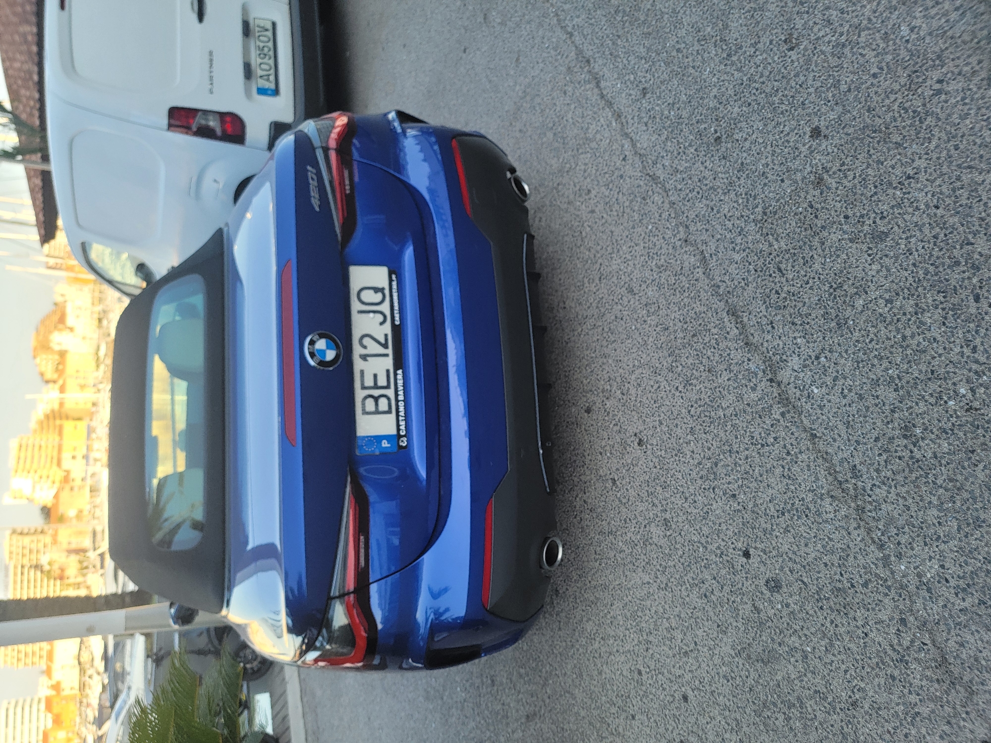 A blue BMW convertible is parked on an asphalt surface in Quarteira, Portugal. The license plate reads "BE12 JQ". Behind the BMW, a white panel van is visible, with a license plate that reads "AO95OV". Buildings with light-colored facades and yellow rooftops are in the background. The scene appears to be a sunny day, with sunlight casting reflections on the vehicles and buildings.