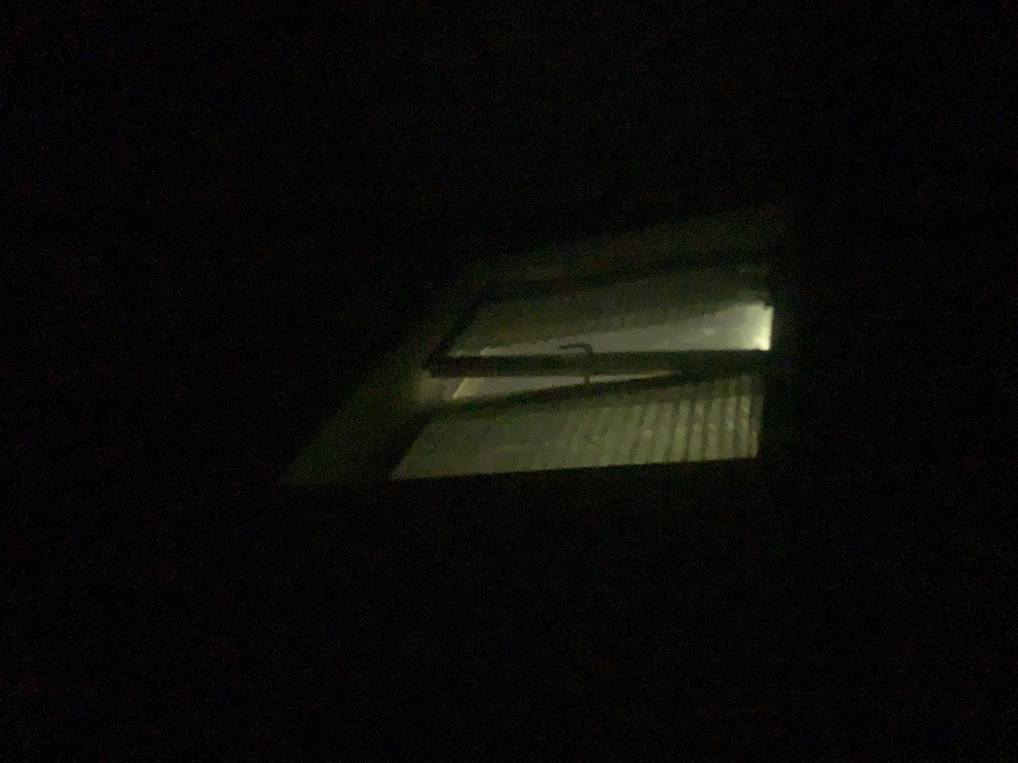 The image shows a dimly lit window at night, likely a residential building. The window appears to have blinds or shutters that are partially open, allowing some light from inside to escape. The surrounding area is dark, suggesting it is nighttime. There are no people or any other distinguishing features visible in the image that would indicate a specific location in Minsk, Belarus.