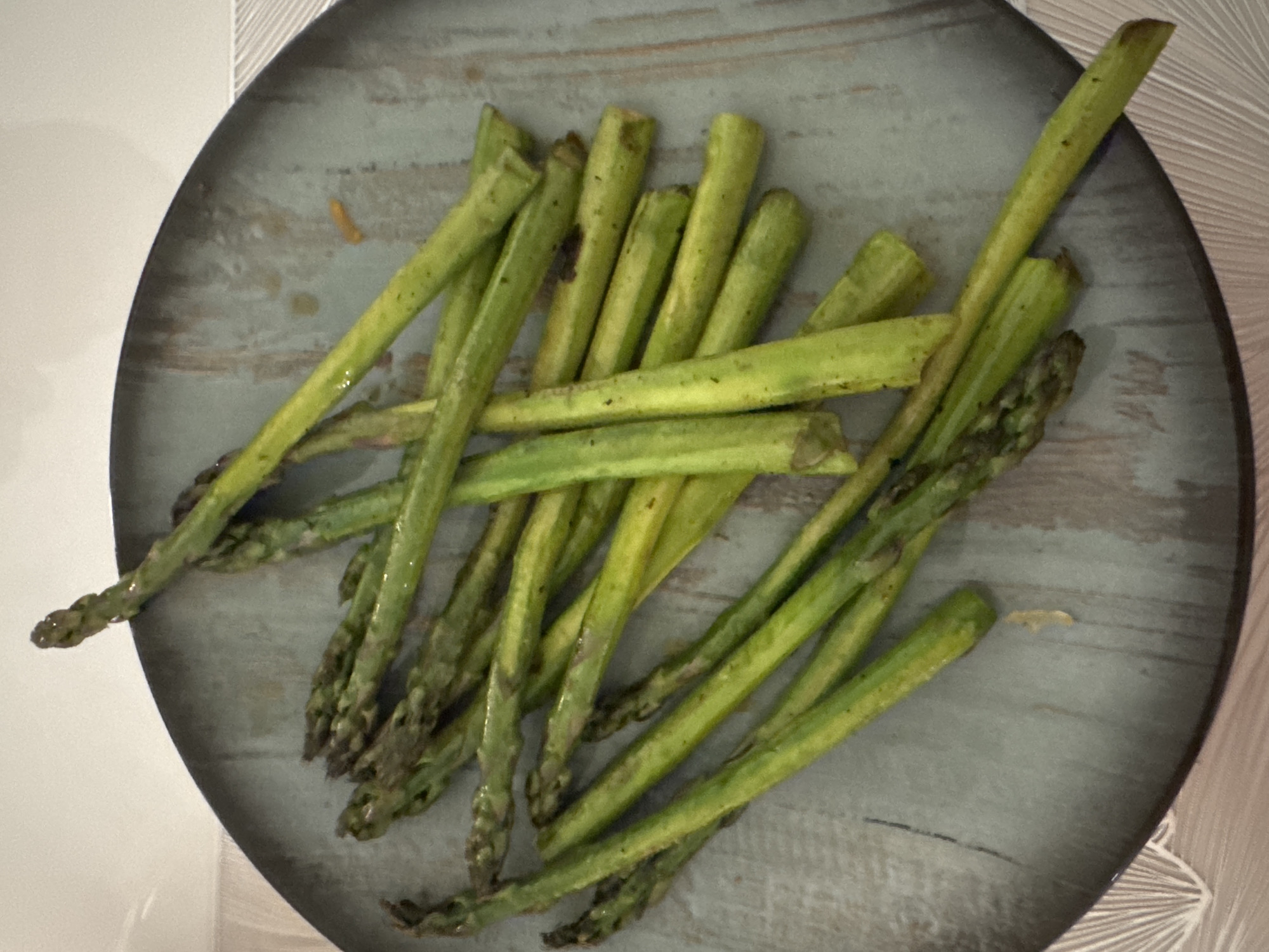 A plate of cooked asparagus is arranged on a round, bluish-gray plate with a slightly distressed finish. The asparagus spears are varying shades of green, some with visible tips and others cut into smaller sections. The plate sits on a surface with a subtle pattern, possibly a tablecloth or placemat. The overall setting appears to be indoors. There is no evidence to suggest the location is in Sofia, Bulgaria.