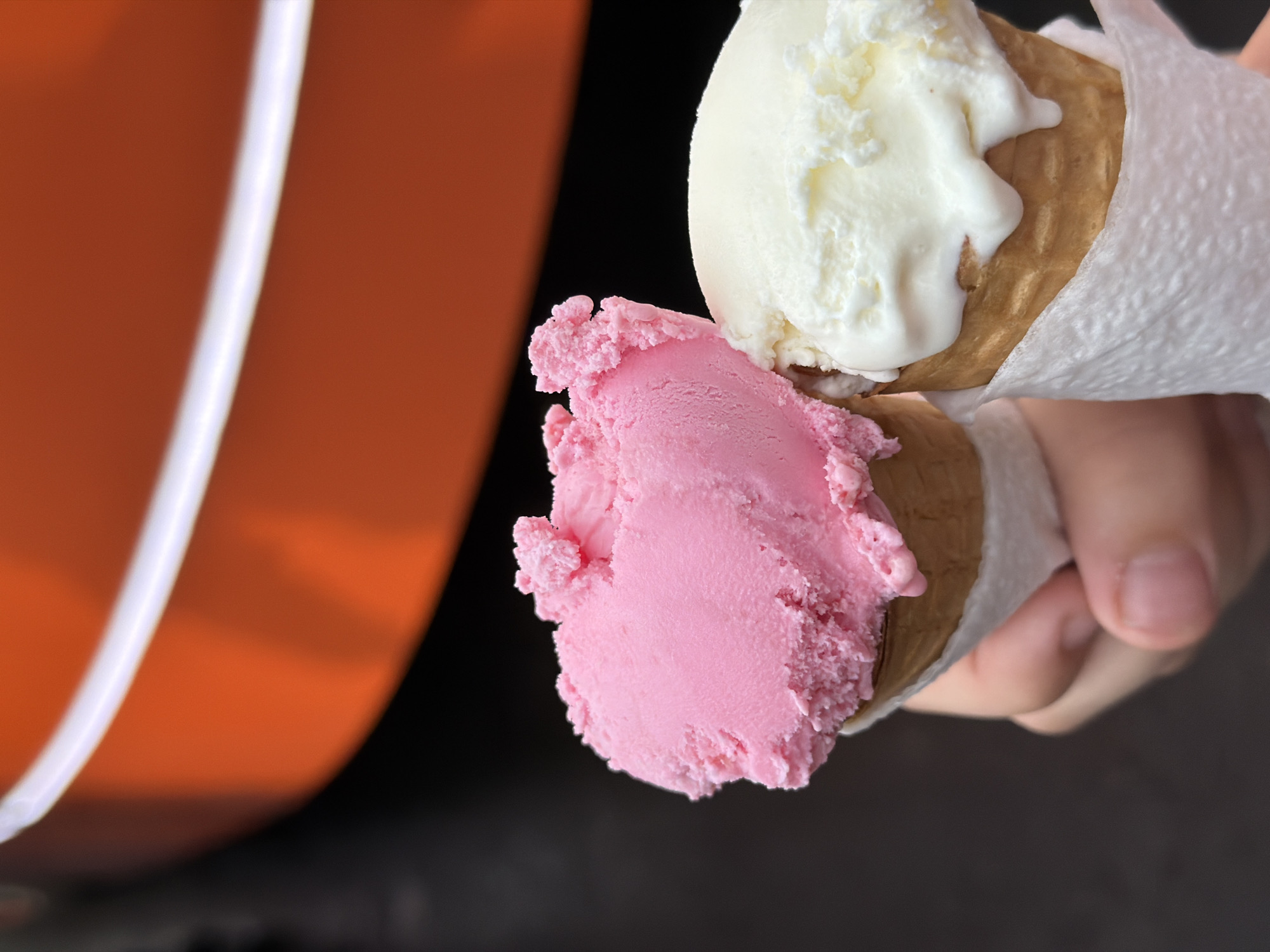 The image shows a close-up view of two ice cream cones being held by a person's hands. One cone is filled with a scoop of pink ice cream, and the other has a scoop of white ice cream. Both cones appear to be waffle cones and are partially wrapped in white paper napkins. The ice cream is slightly melting, indicating it is a warm day or that the ice cream has been out for a short period.
In the background, there is a large, out-of-focus object that appears to be orange with a white stripe. This could be a part of a building, a vehicle, or some other large structure. The rest of the background is dark and out of focus, providing a neutral contrast to the brightly colored ice cream.
The image is well-lit, with the light source highlighting the texture and details of the ice cream and cones. The focus is on the ice cream, suggesting that the primary subject is the food itself. The way the cones are held implies someone is about to enjoy them. There are no discernible emotions conveyed, but the presence of ice cream often evokes feelings of enjoyment and refreshment. No text is visible in the image. The scene appears to be outdoors or in a bright indoor setting, given the lighting and the context of ice cream.
